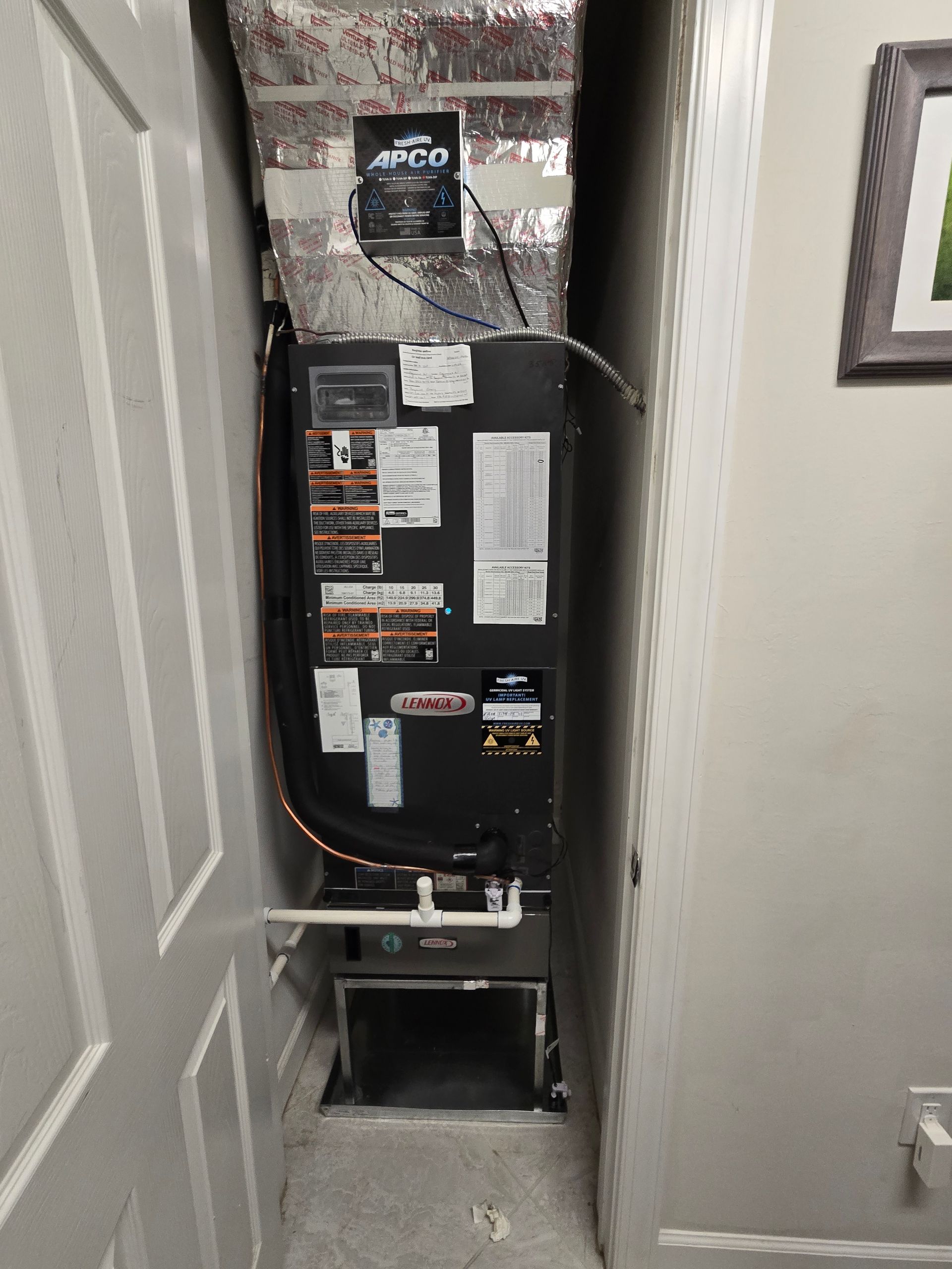 Furnace unit in a narrow utility closet, with ductwork and service labels. Gray walls and a white door frame.