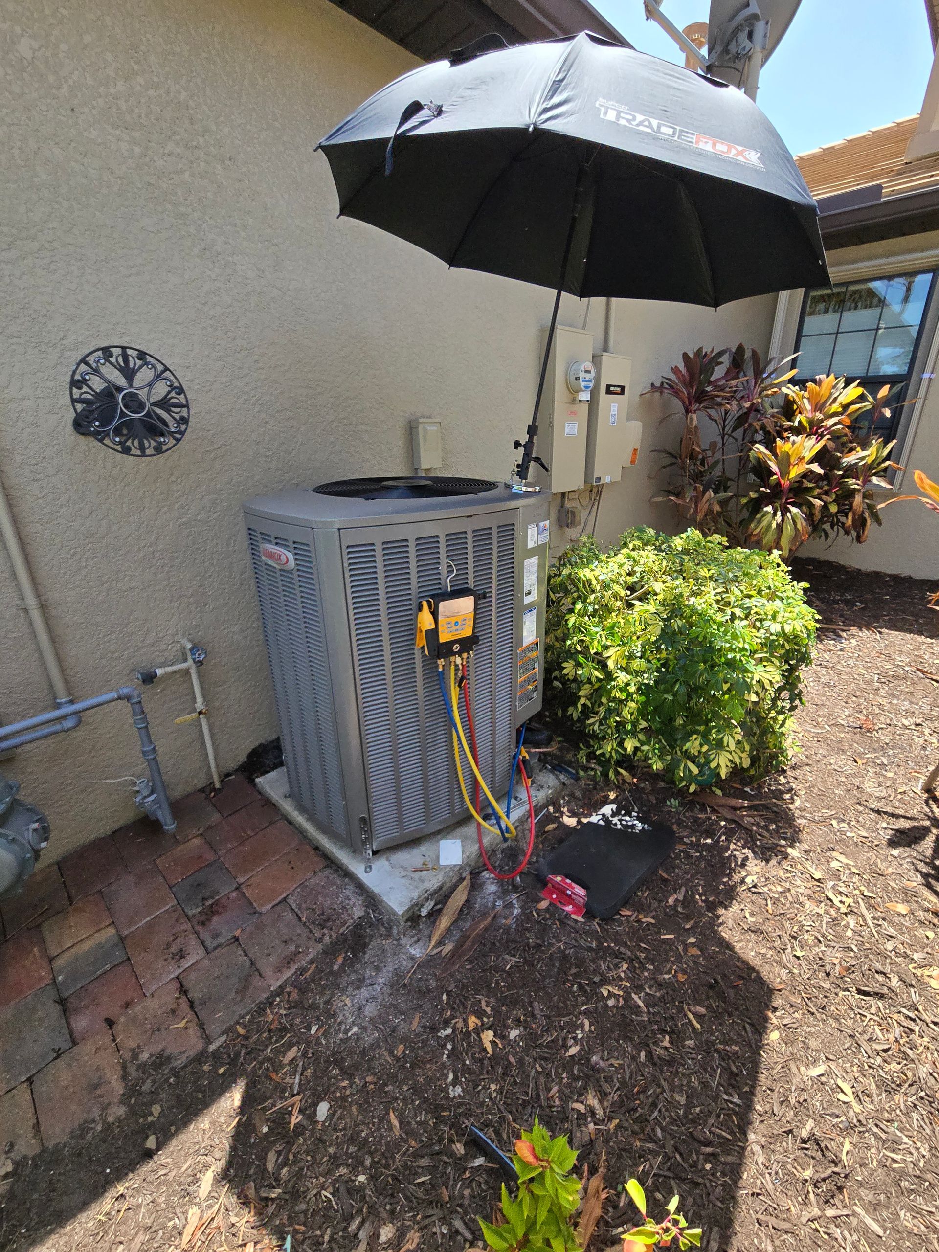 HVAC unit being serviced outdoors; a black umbrella provides shade. Tools and gauges are connected.