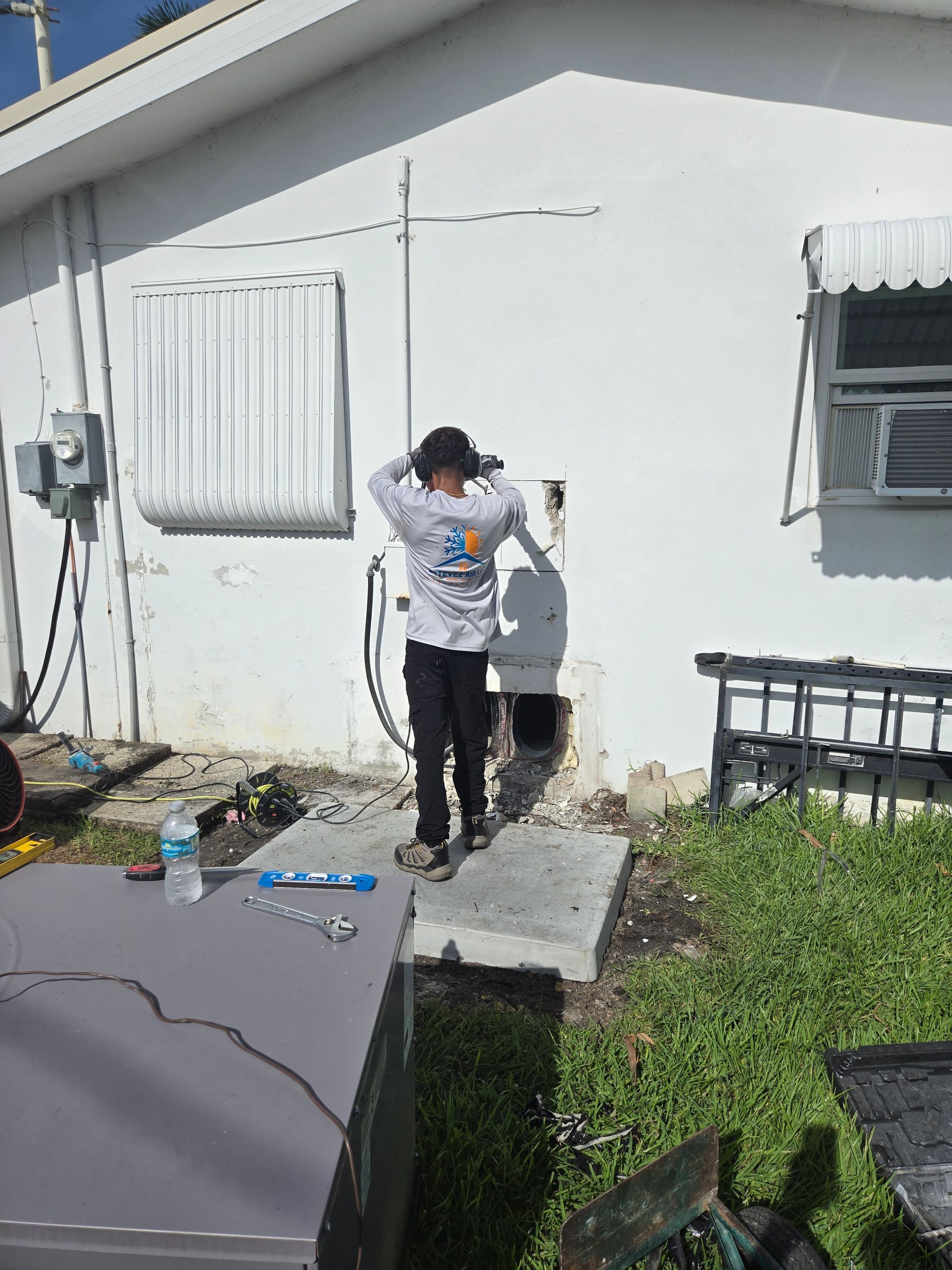 Person adjusting something outside a white building. Tools and a metal object are in front.