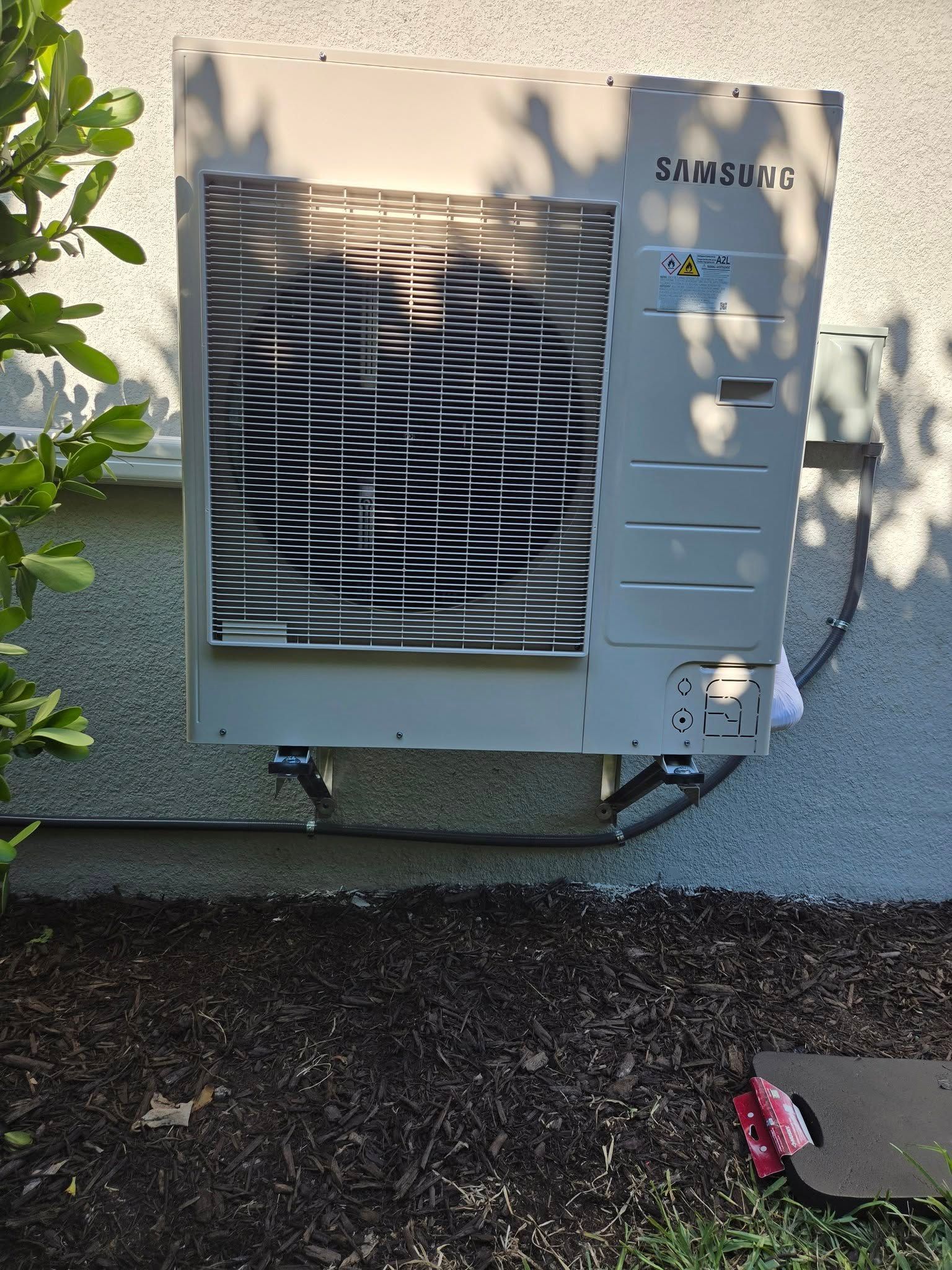 Samsung air conditioning unit mounted on a wall above mulch, near green foliage.