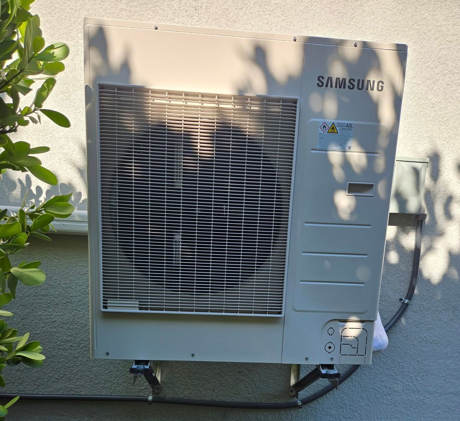 Samsung air conditioning unit mounted on a wall above mulch, near green foliage.