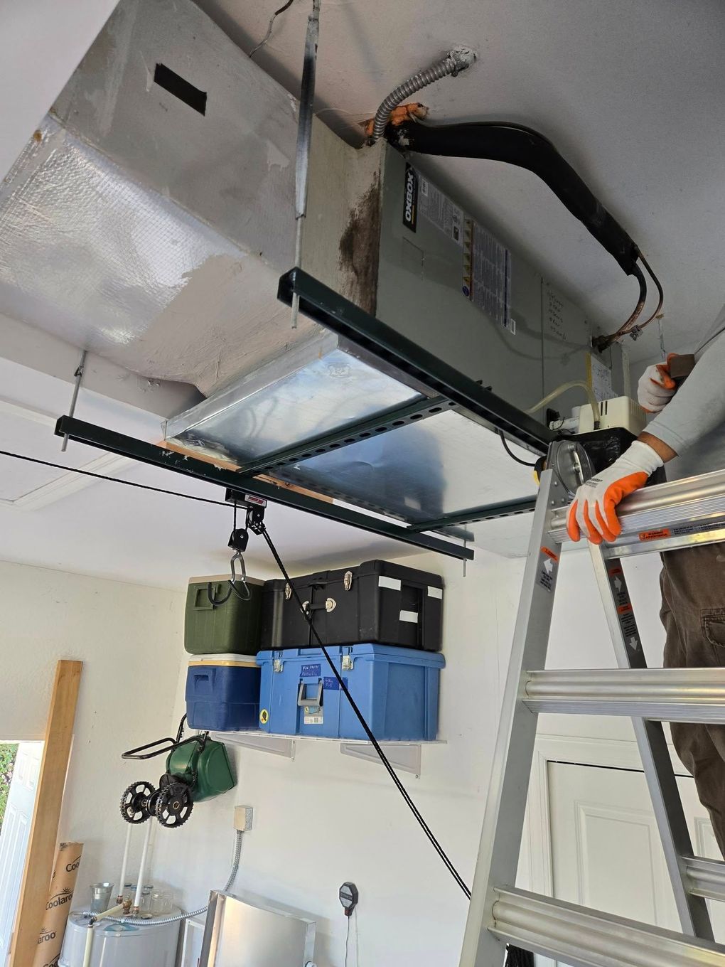 Person on a ladder near an HVAC unit, installed on a garage ceiling, with storage bins below.