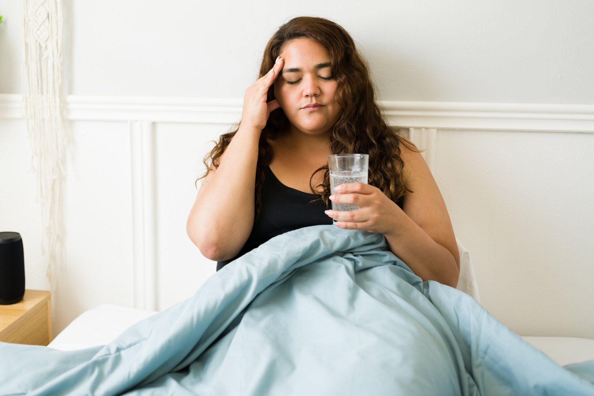 young hispanic woman with headache in bed holding glass of water