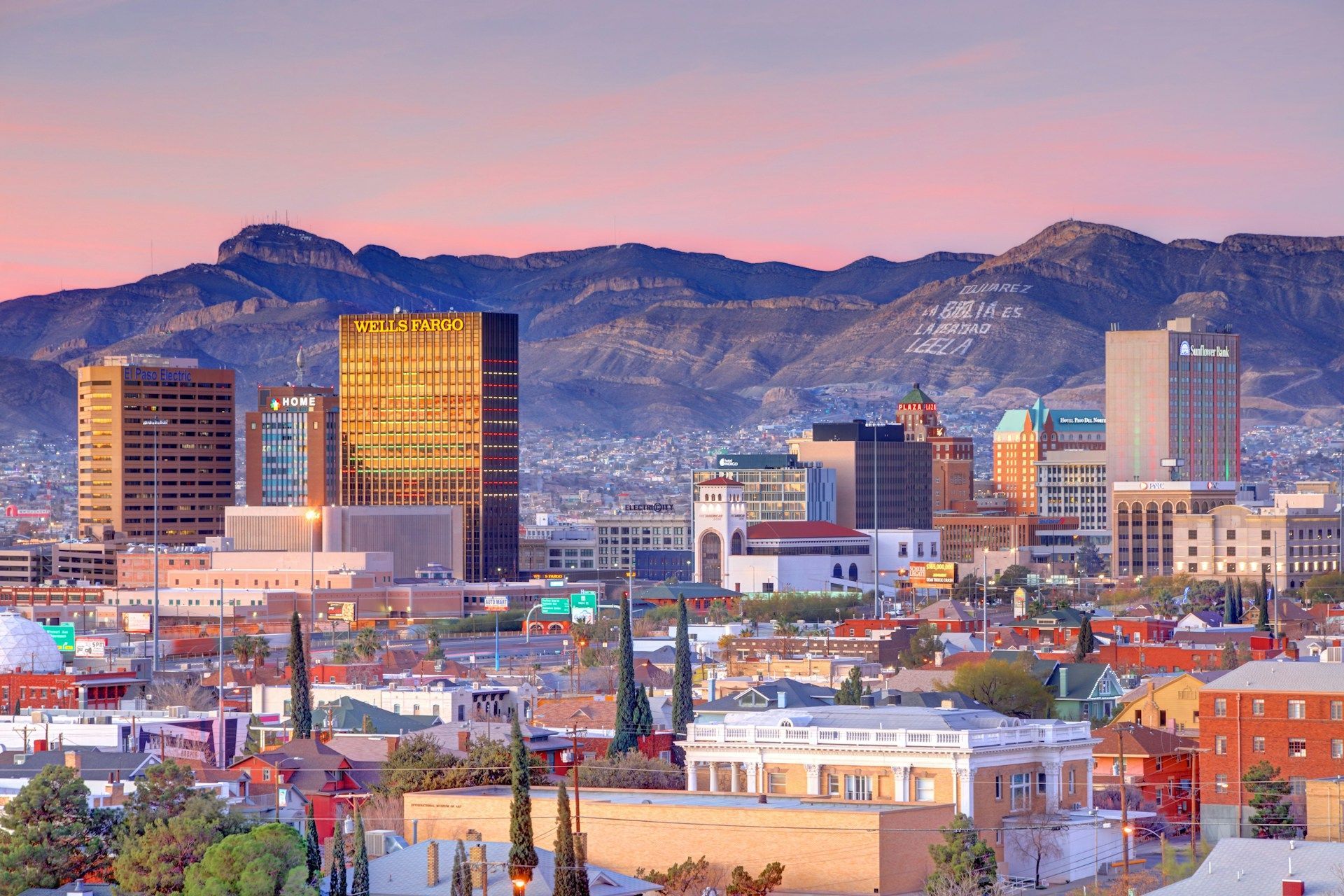 City skyline with buildings, mountains in the background, under a pink and orange sky.