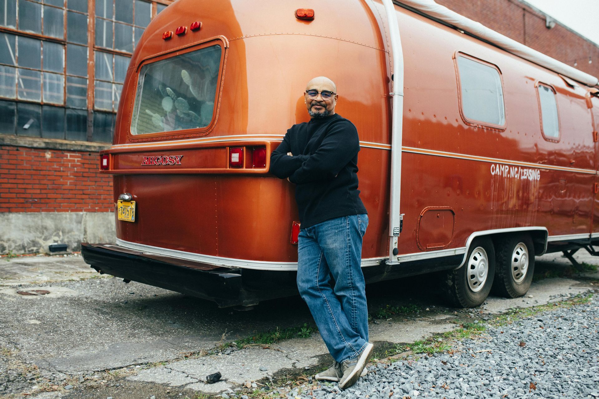 Man in jeans leans against an orange travel trailer, arms crossed. Outdoors, brick building backdrop.