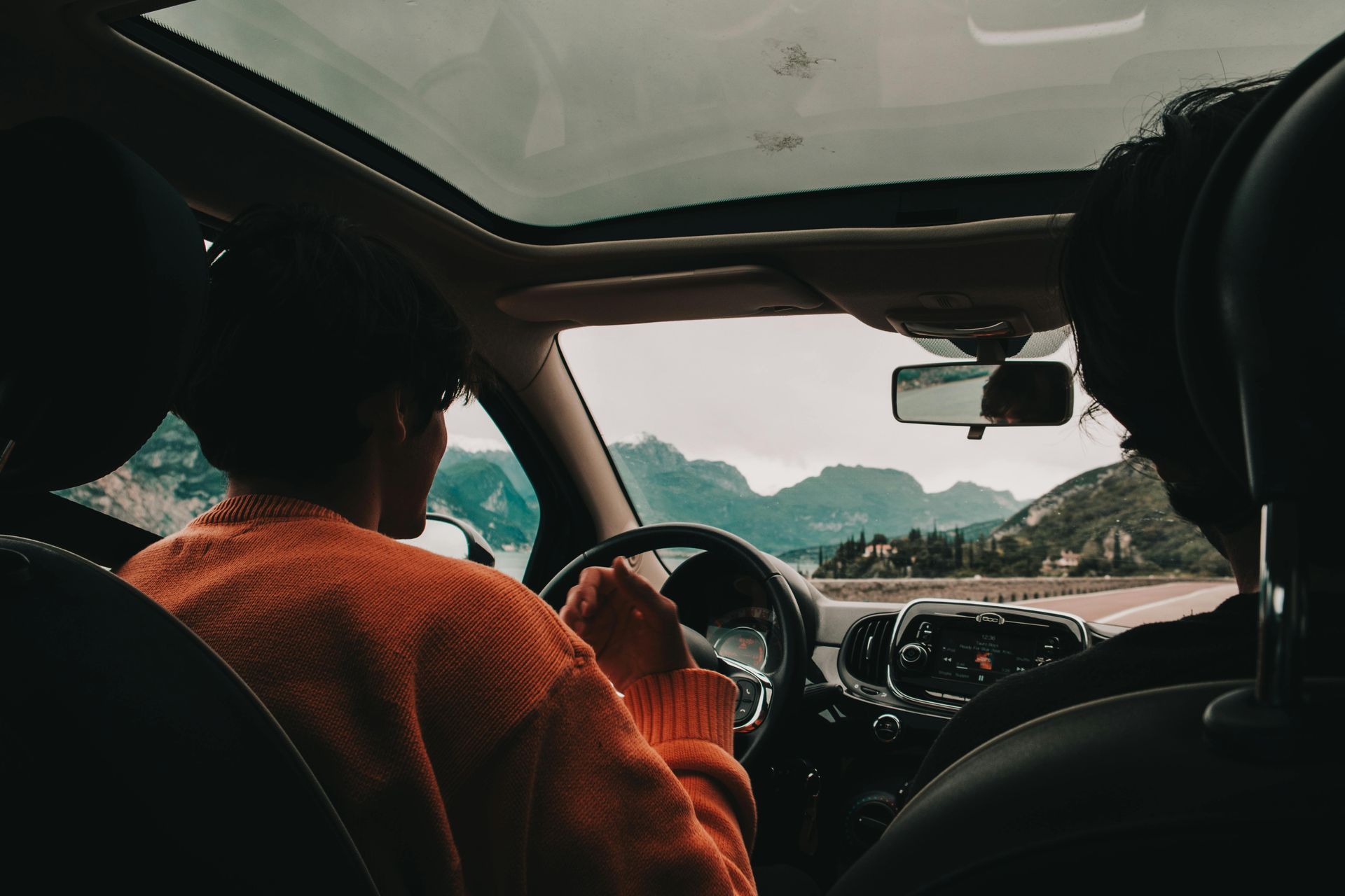 Person driving car with mountains in the distance, viewed from inside. Skyroof open. Orange sweater.