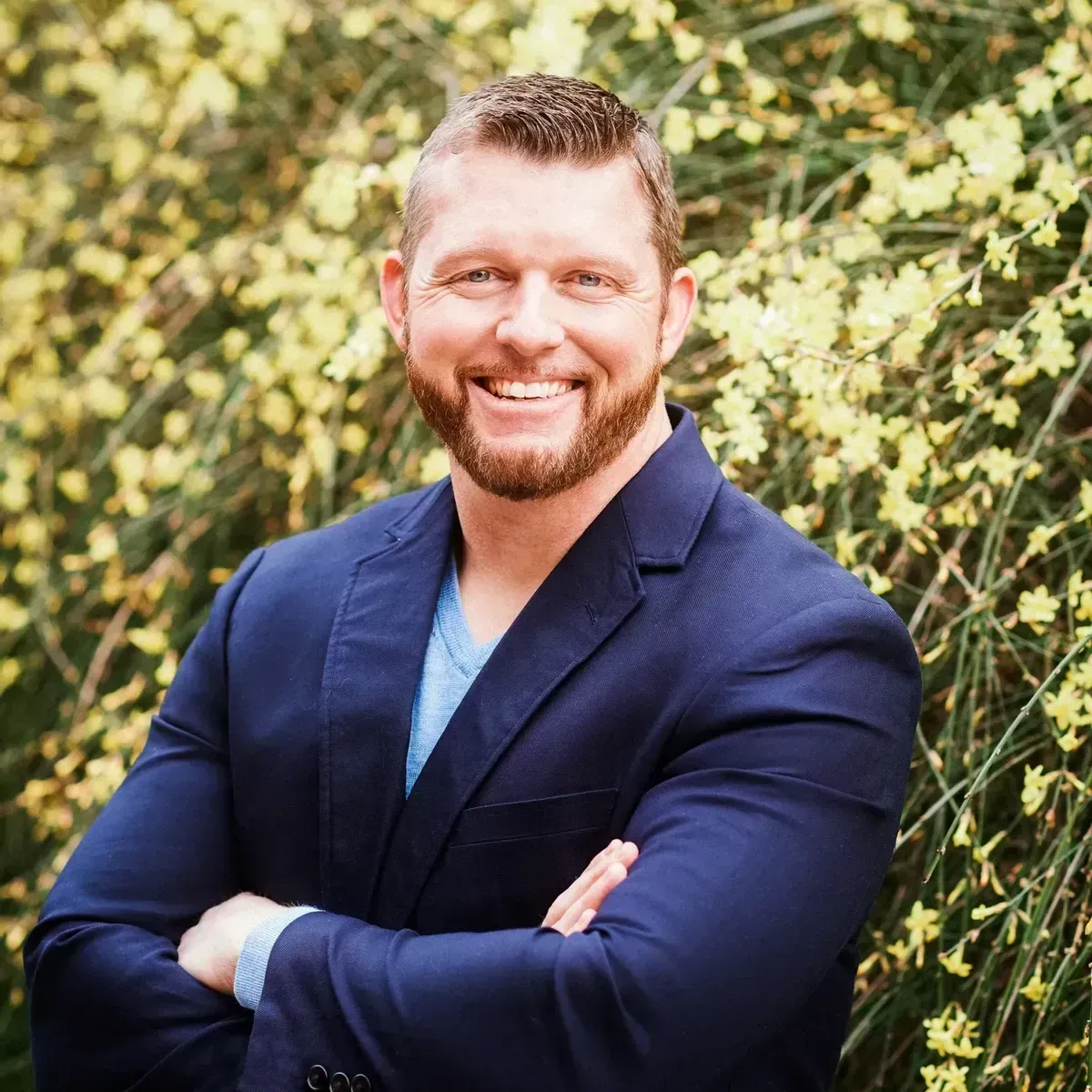 A man in a blue suit is standing in front of a bush with yellow flowers.