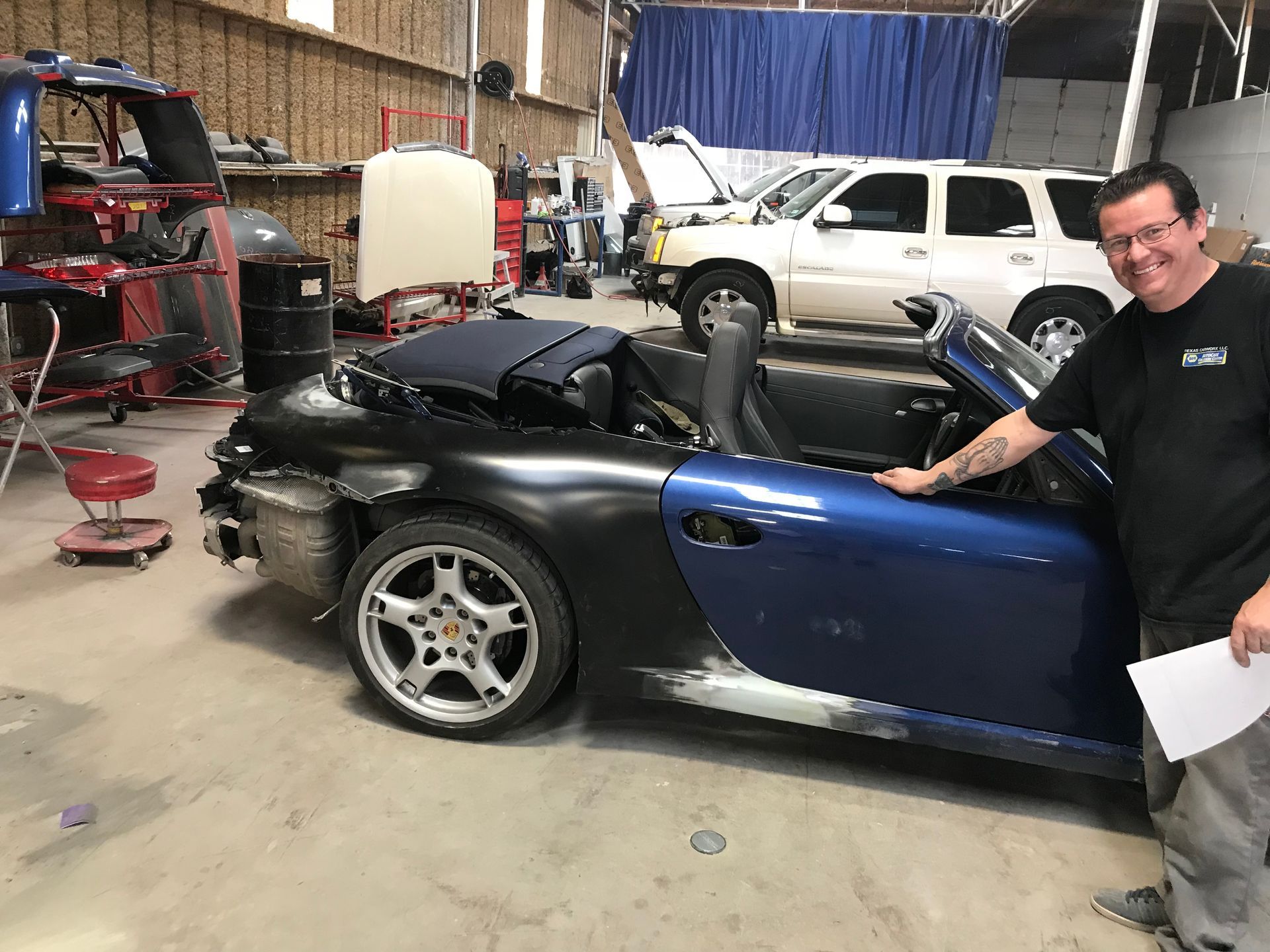 Man smiling beside a black-and-blue convertible in a garage, with other cars and tools in the background | Texas Carworx