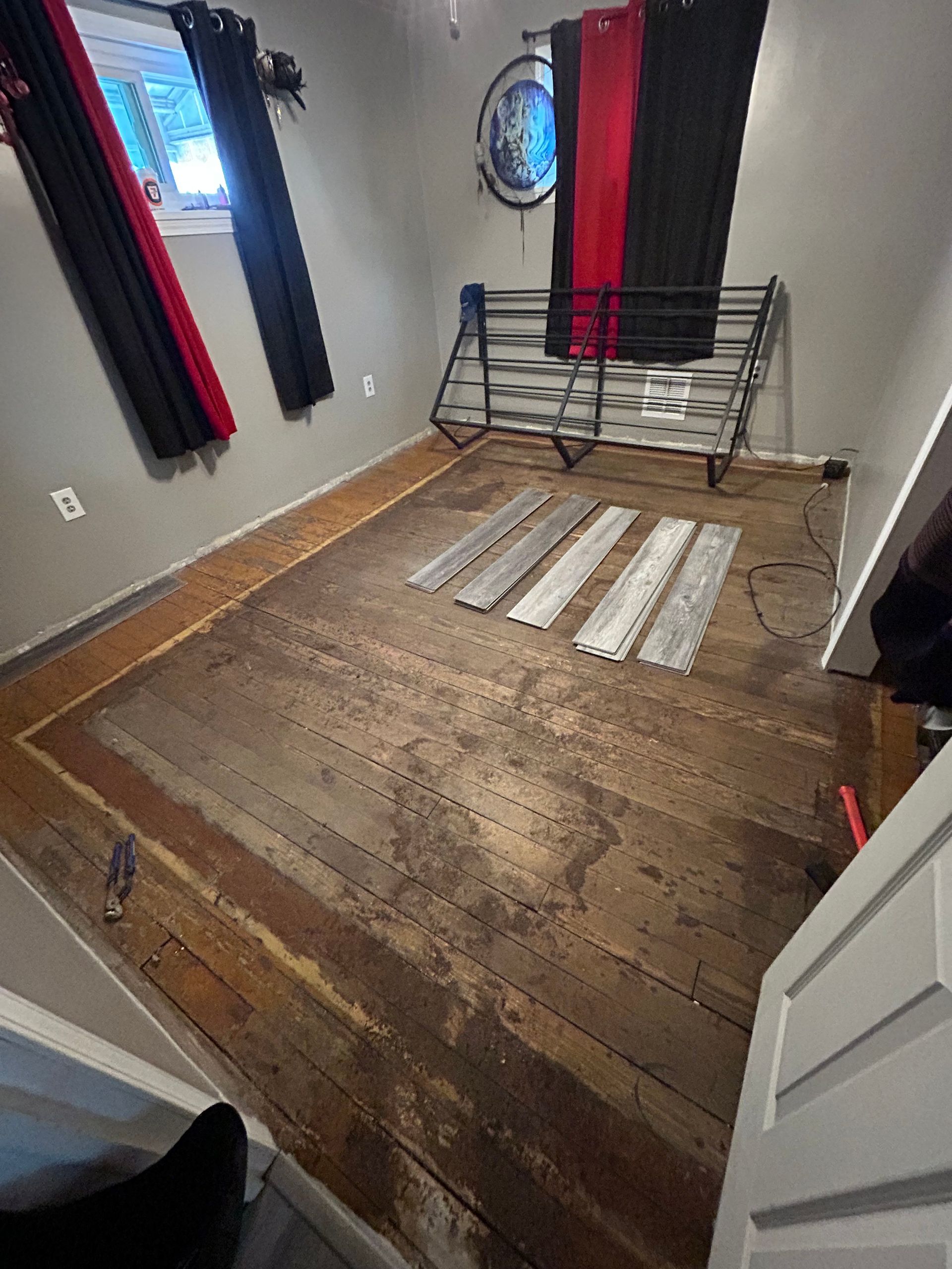 A room with damaged, worn wooden floor and a bed frame. Window with curtains, exposed wiring.