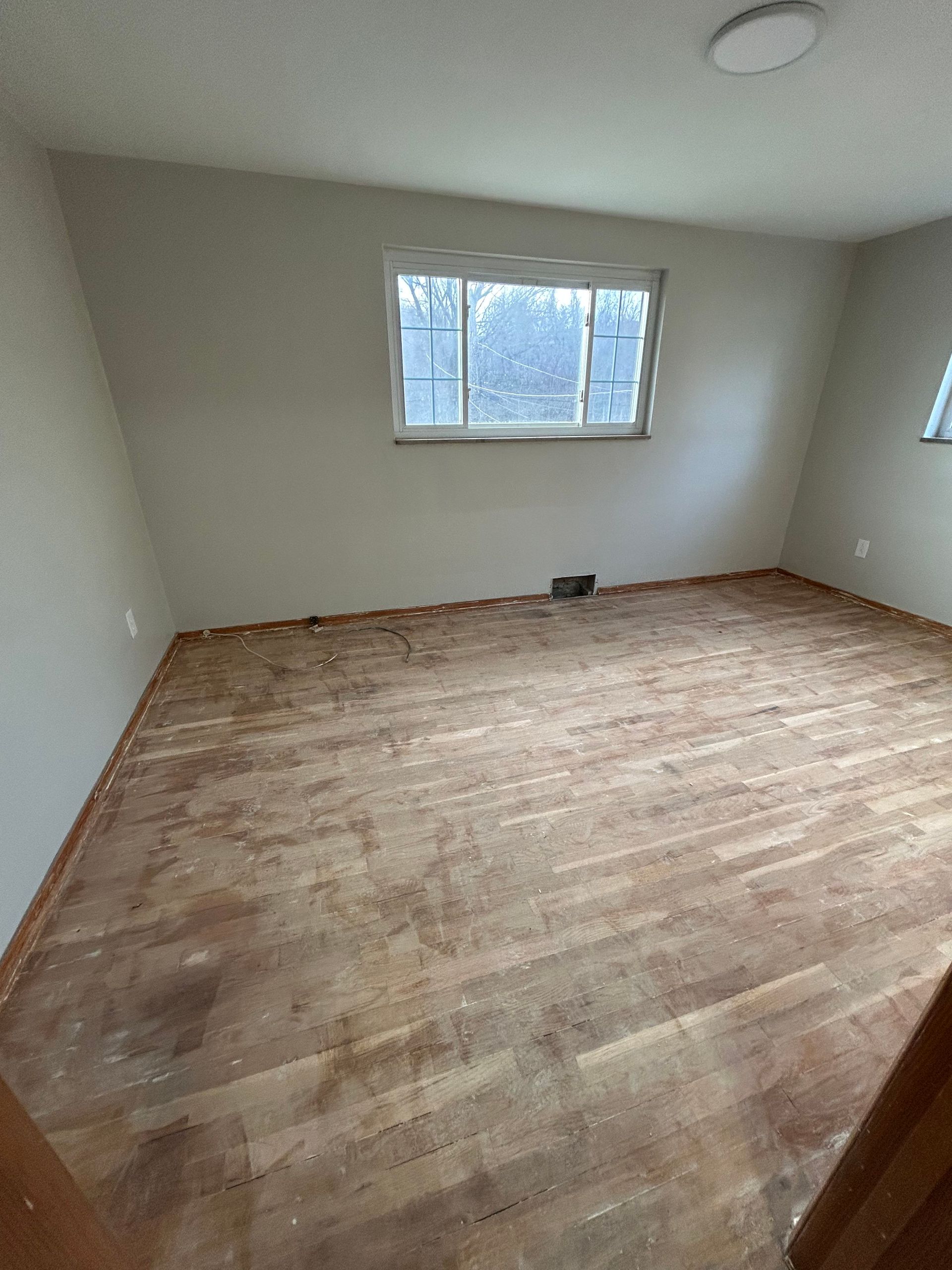 Empty room with light walls and worn wooden floor. A window is centered on the wall.