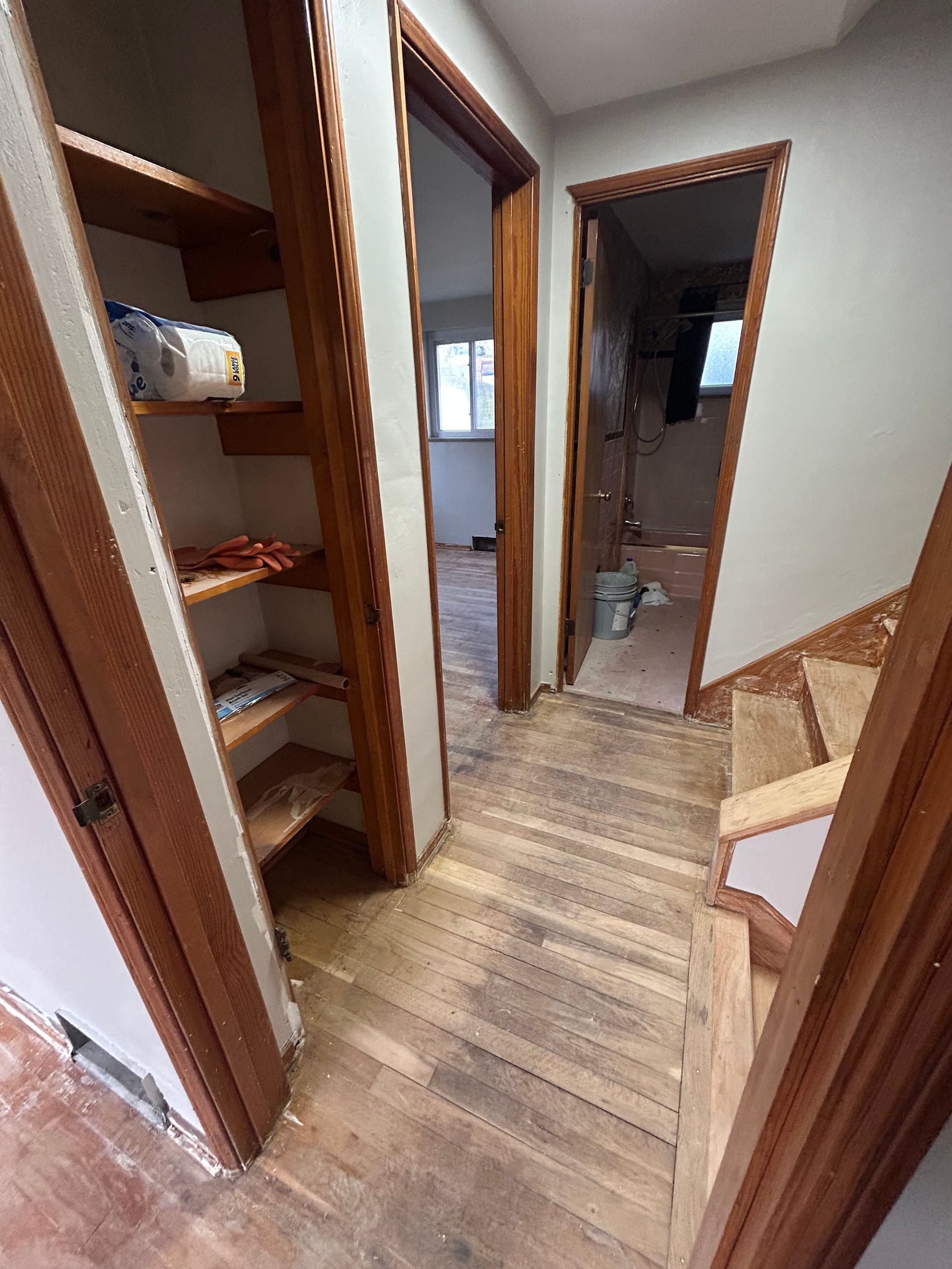 Hallway with stairs and doorways. Wooden stairs and trim, white walls. One doorway leads to storage shelves.