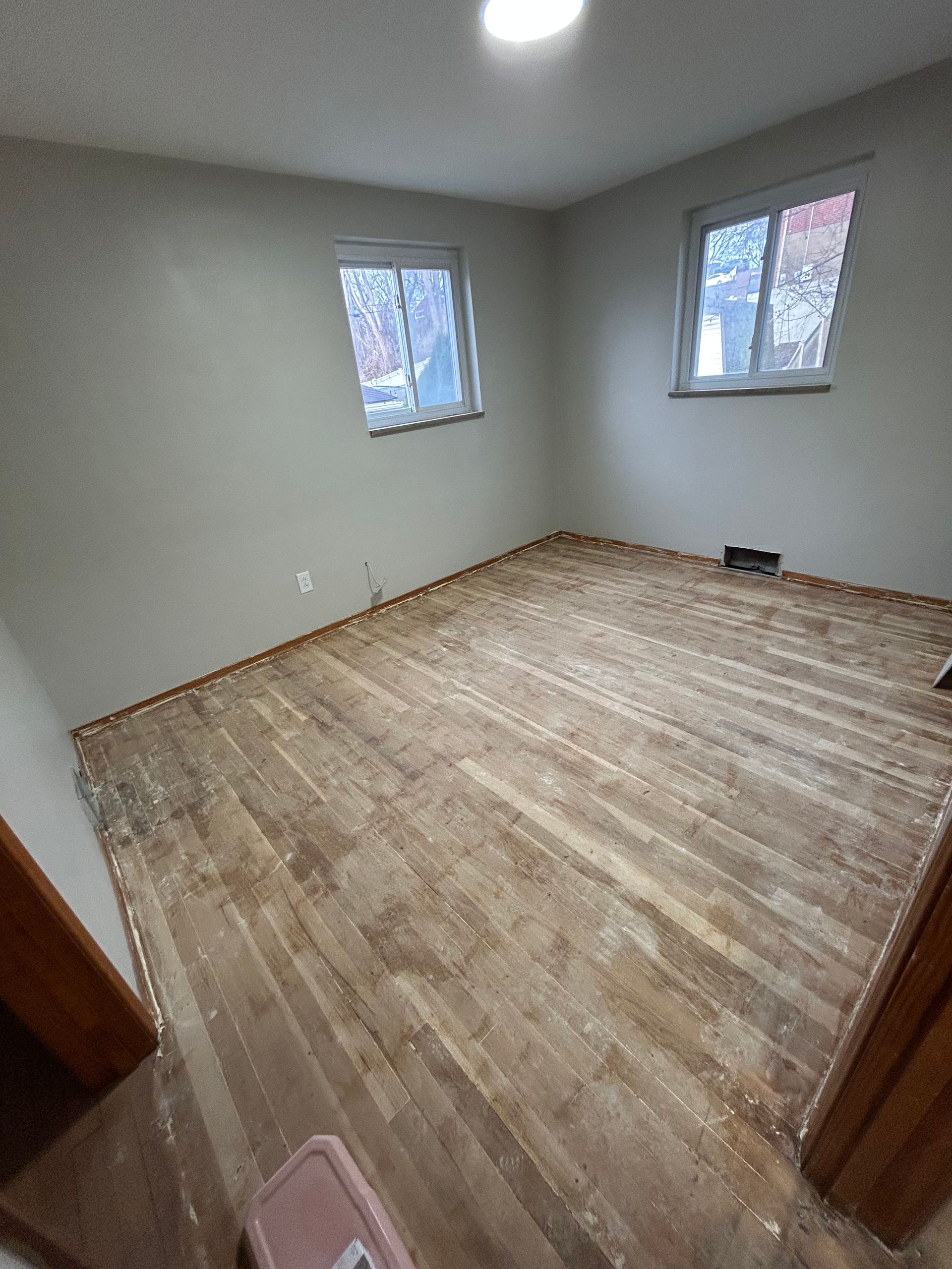 Empty room with worn wood flooring and two windows, neutral walls, overhead light.