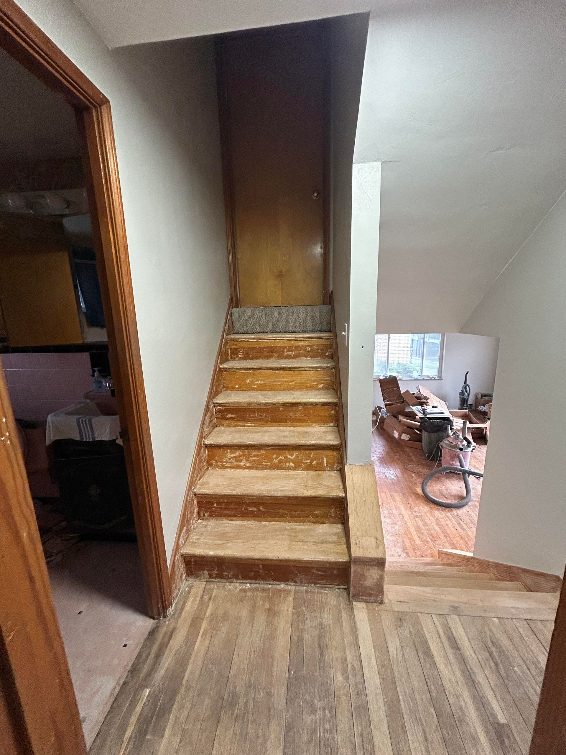Wooden staircase leading up to a closed door, with a hallway and living area visible.