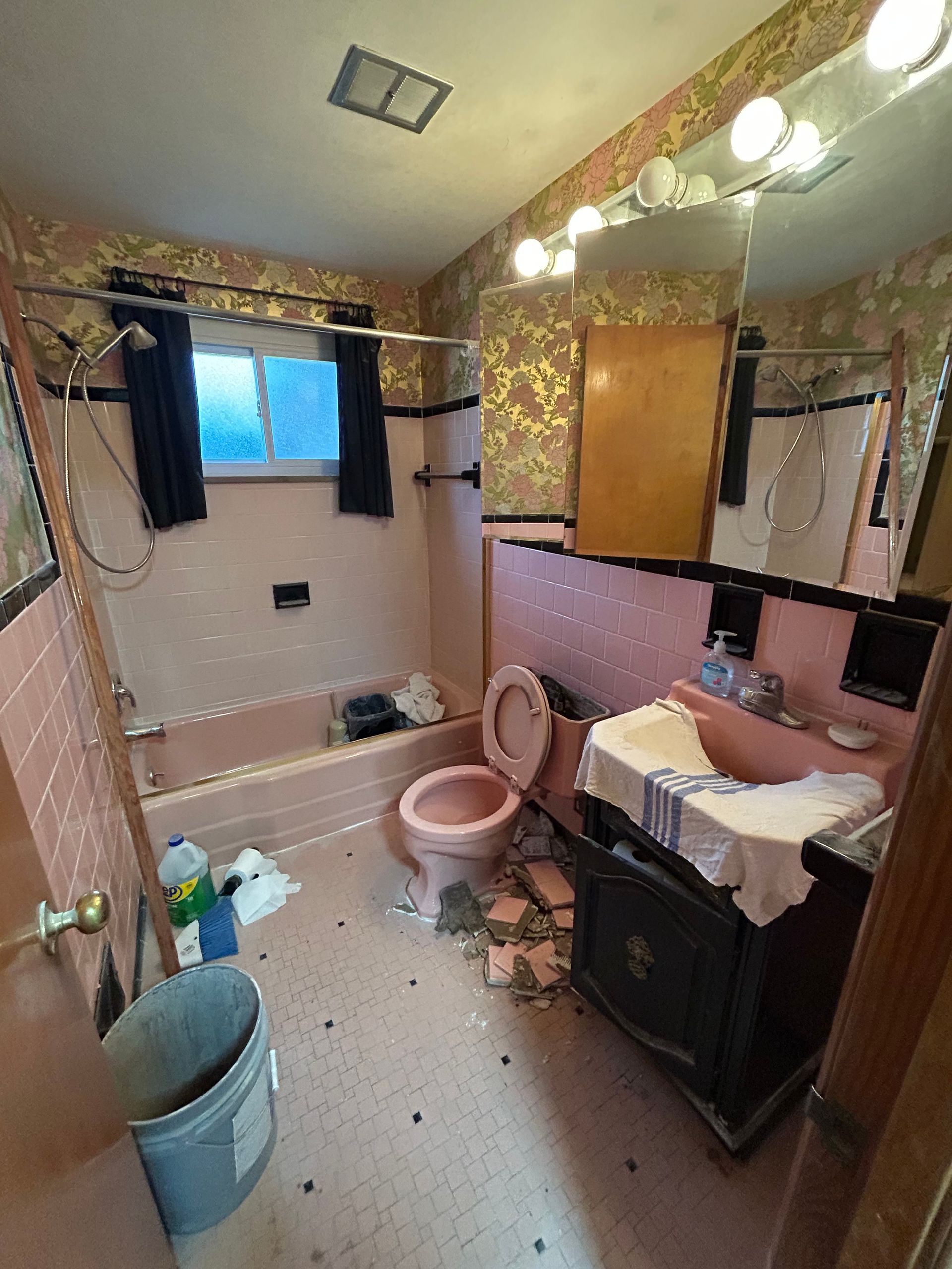 Pink and black bathroom in disrepair: pink fixtures, gold wallpaper, debris on the floor, and a bucket.