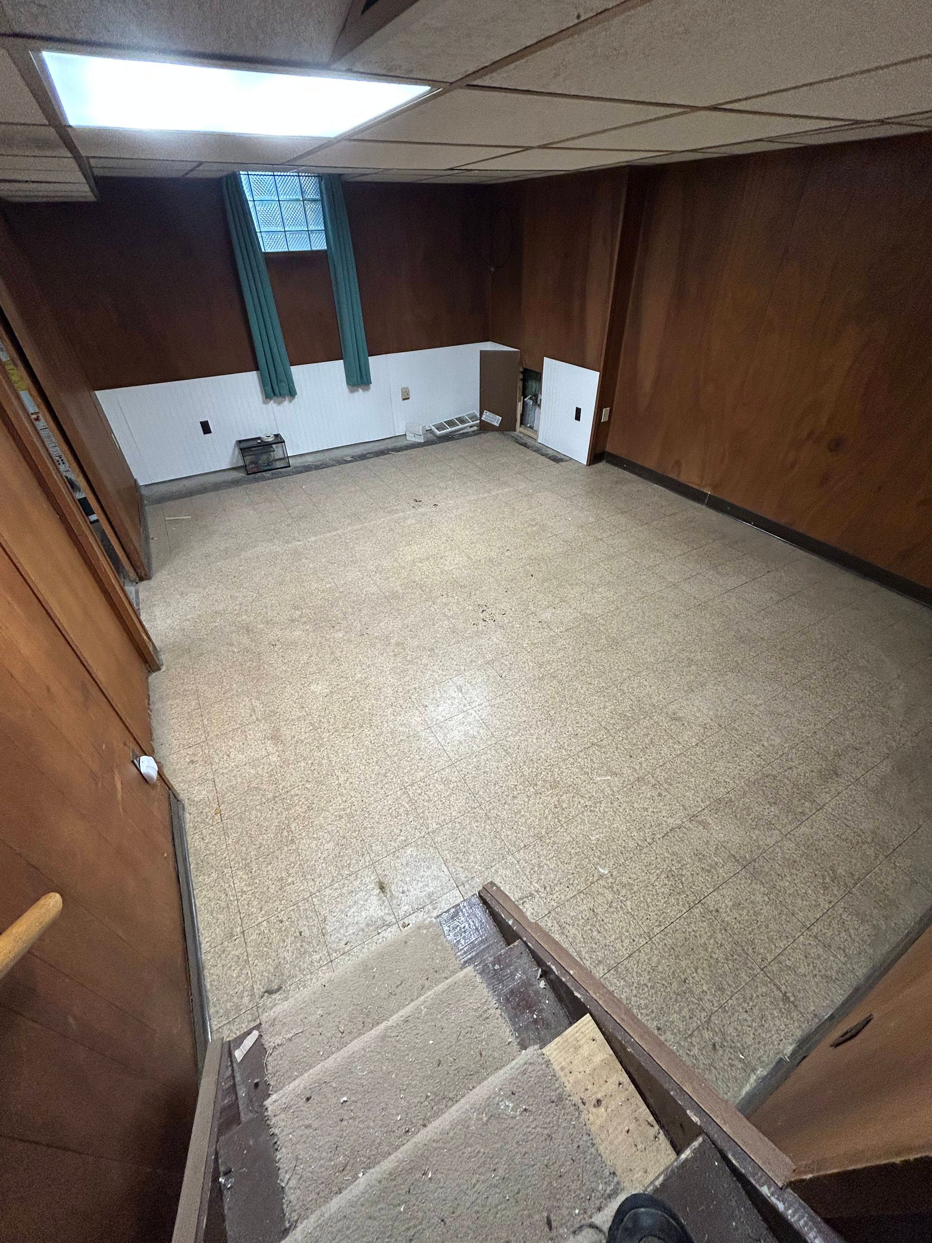 Basement room with wood-paneled walls, speckled floor, stairs, and a window.