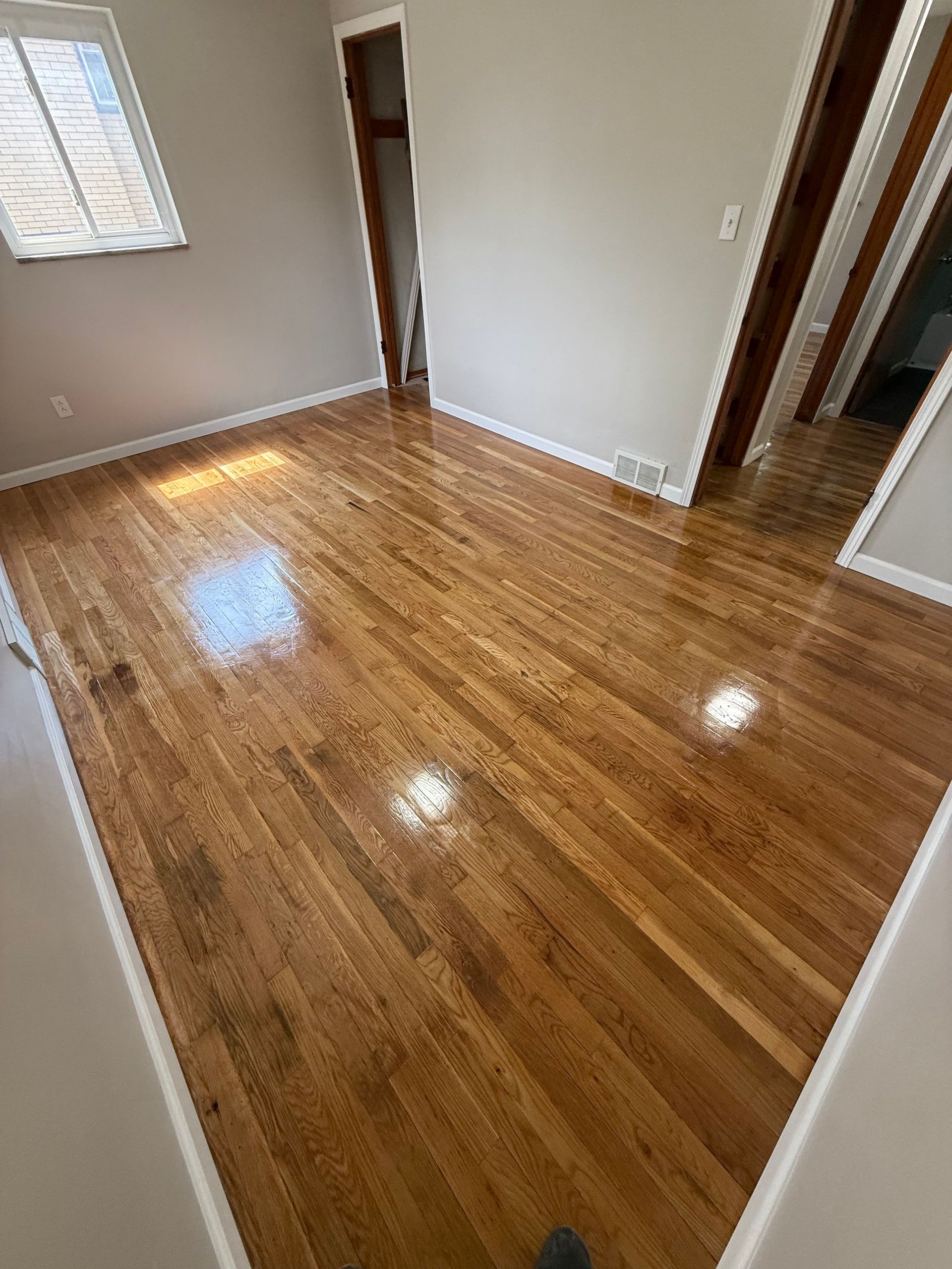 Newly refinished hardwood floors in an empty room, reflecting light. White trim, neutral walls, and a doorway are visible.