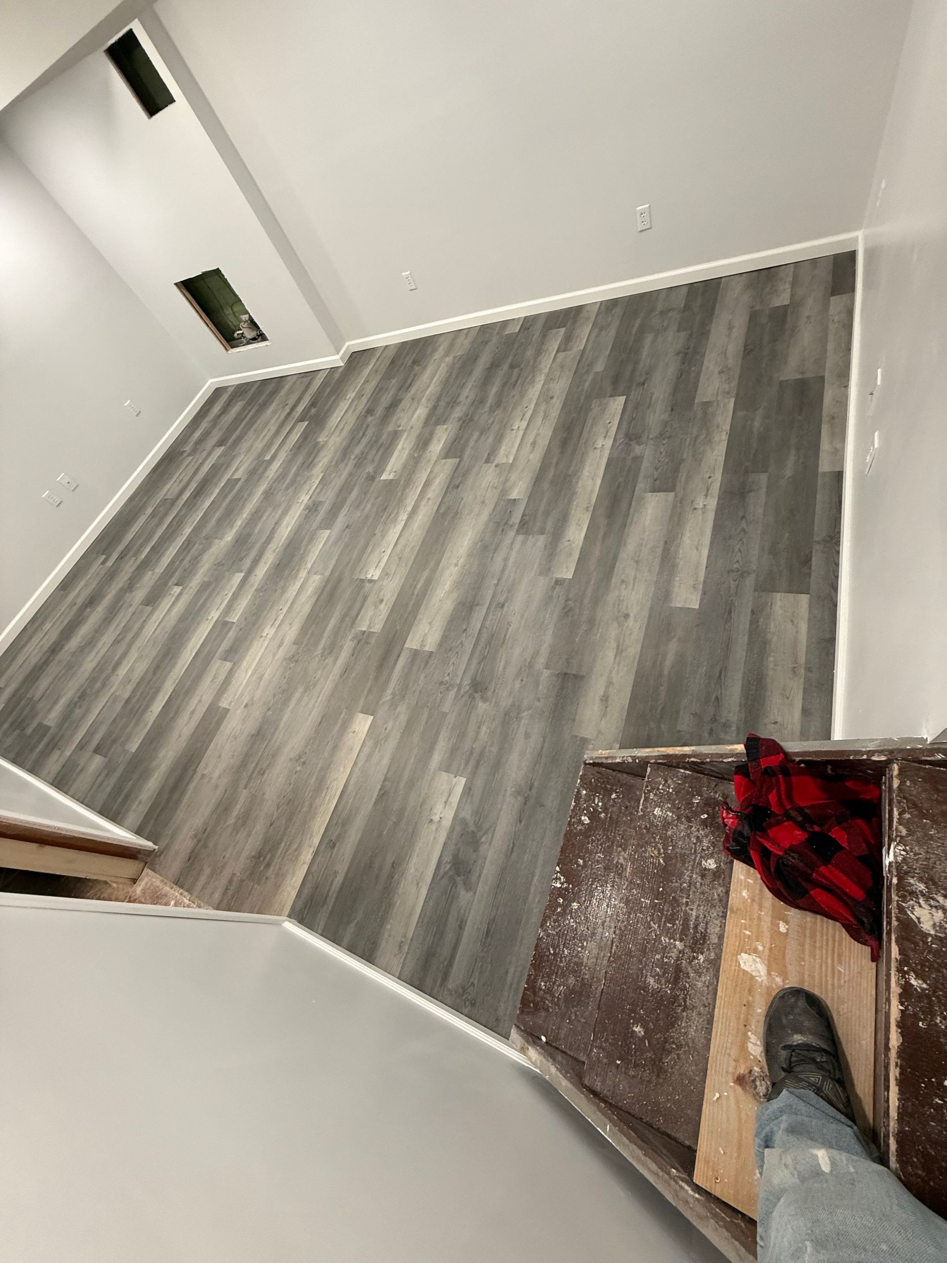 Overhead view of a room with gray wood-look flooring and white trim. Someone's foot and a plaid shirt are visible.
