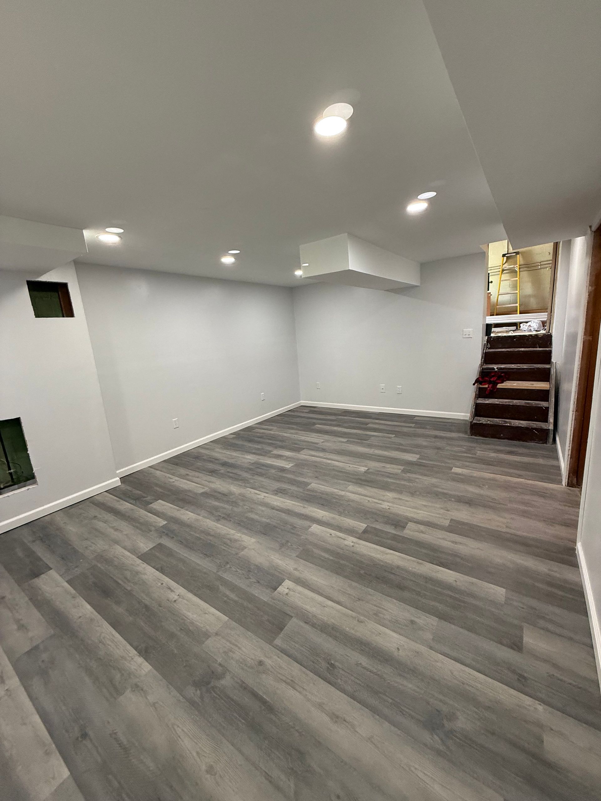 Empty finished basement with gray wood-look flooring, light gray walls, and recessed lighting. Stairs lead up.