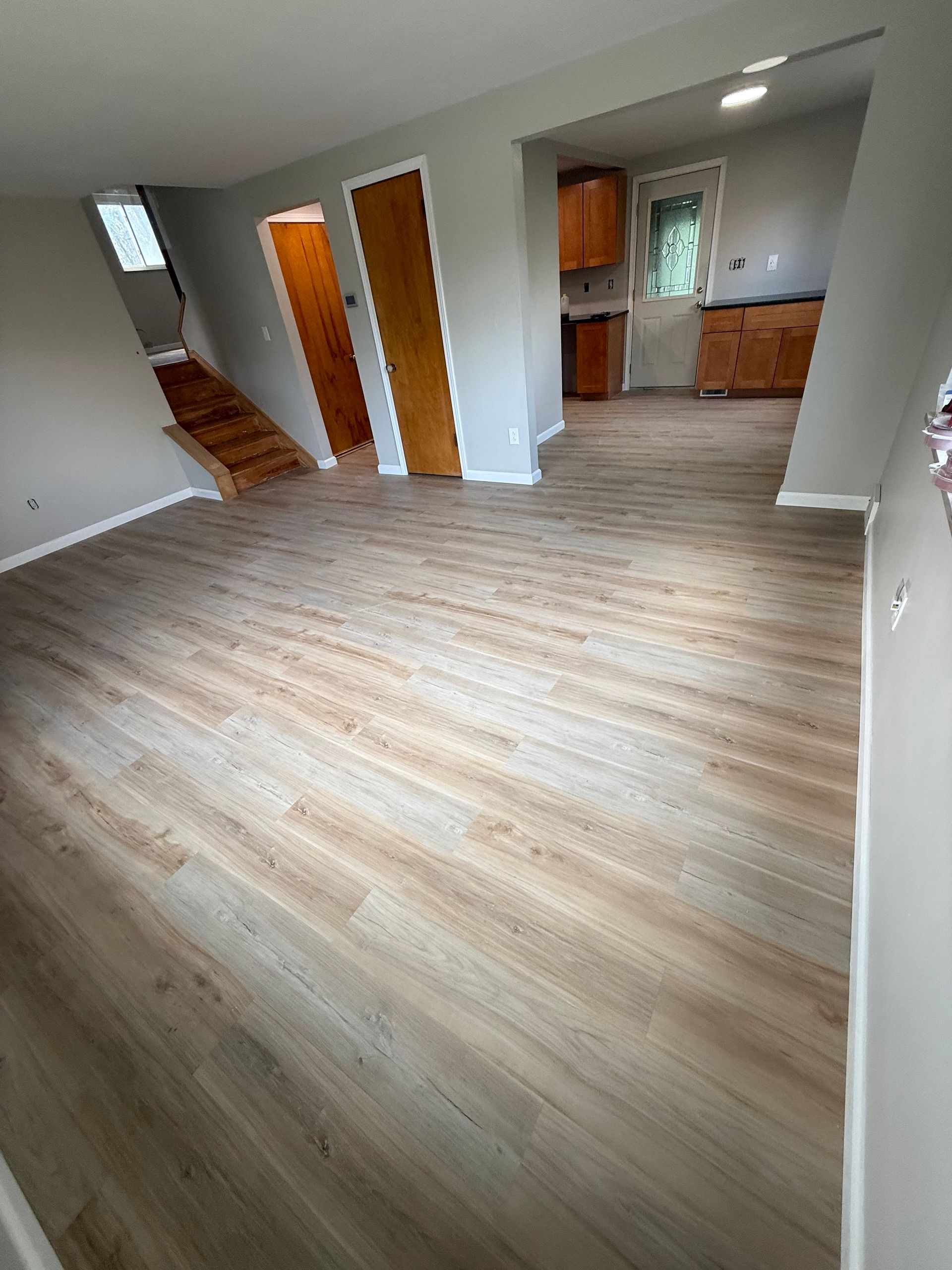 Empty living space with wood-look flooring and gray walls. Kitchen and staircase visible in the background.