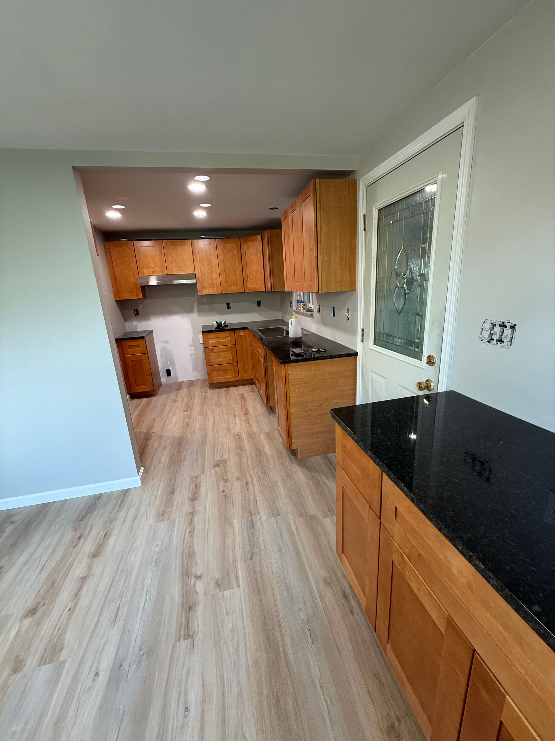 Kitchen with wooden cabinets, black countertops, and light wood flooring. A door is to the right.