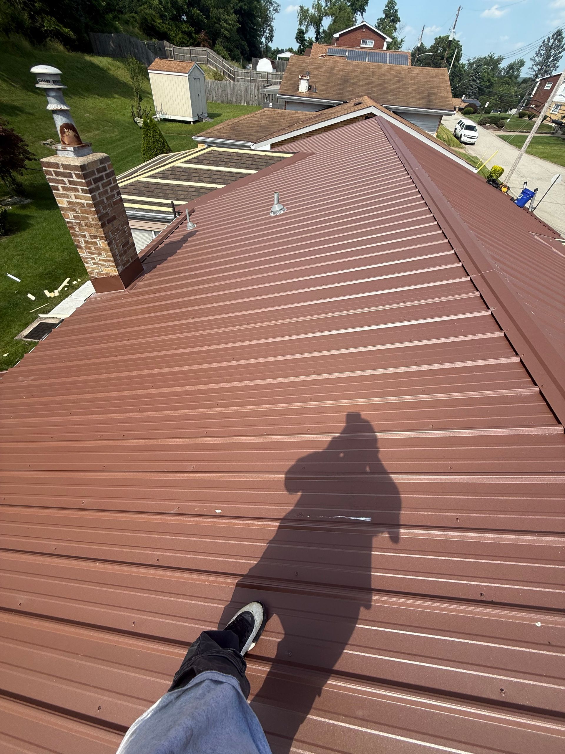 Person's shadow on a brown metal roof. A chimney and houses are in the background. Bright sunny day.
