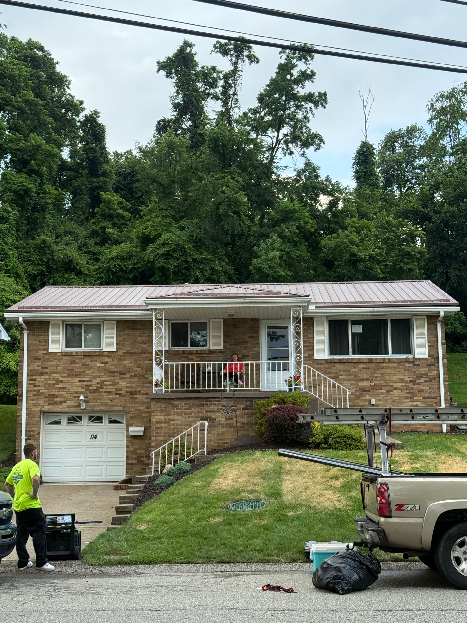 Brick house with red metal roof, driveway, and lawn. Person in yellow shirt. Truck with ladder parked nearby.
