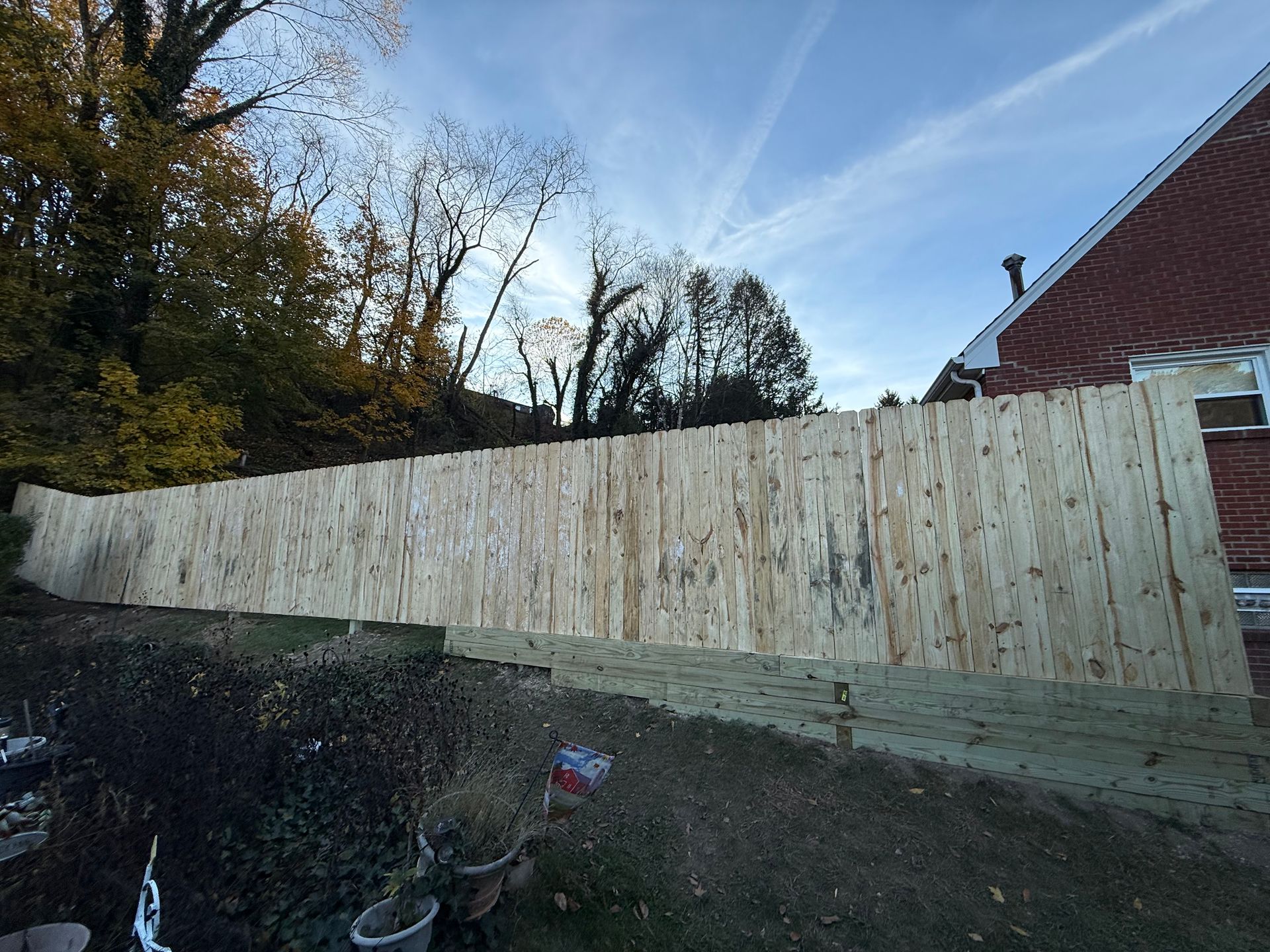 Wooden fence runs along a sloping yard, next to a brick building under a cloudy sky.
