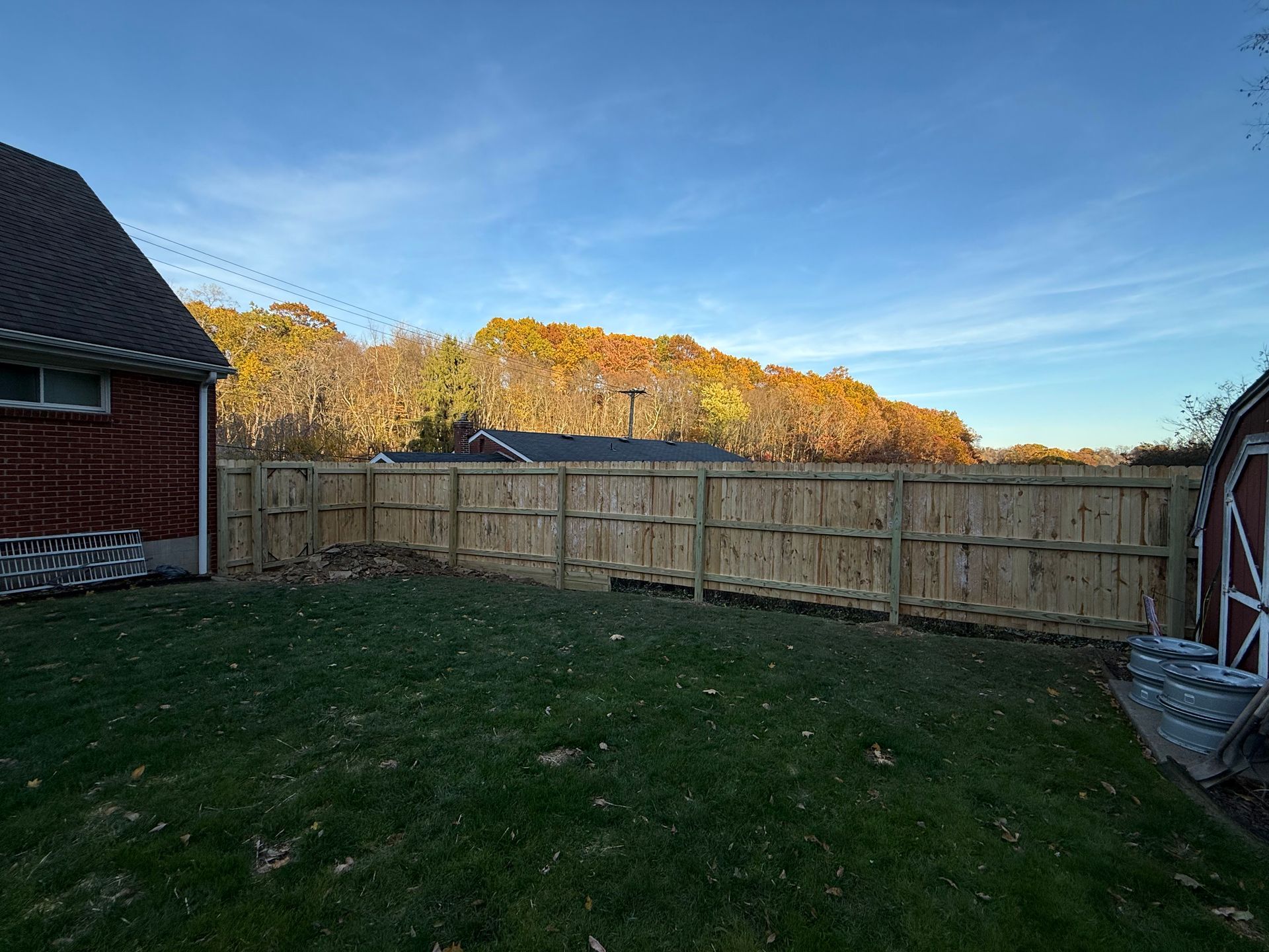 Wooden fence surrounds a green grassy backyard. Red brick building and trees with fall foliage are in the background.
