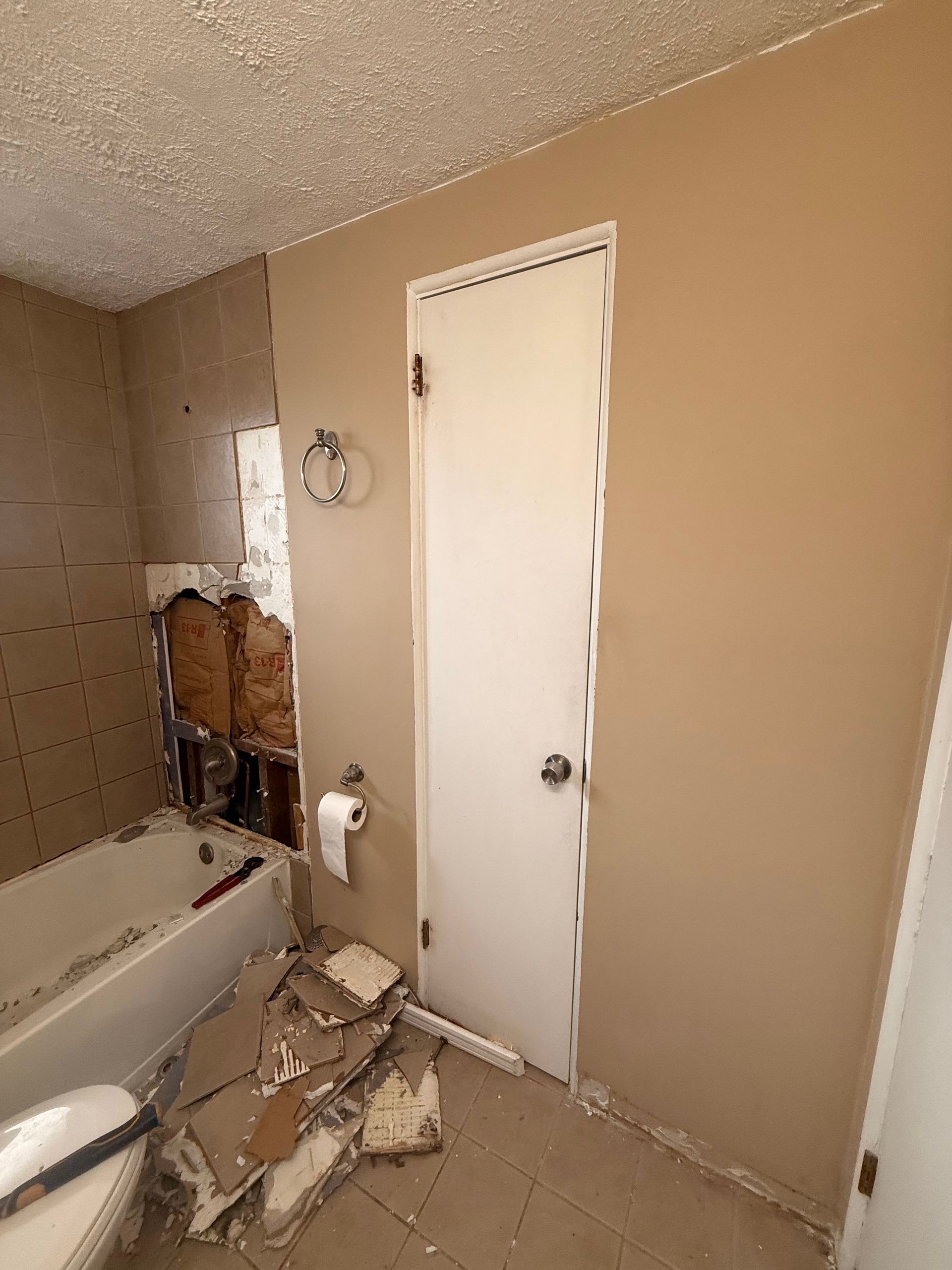 Bathroom renovation, drywall removed around the tub, debris on floor, door to the right. Beige walls.