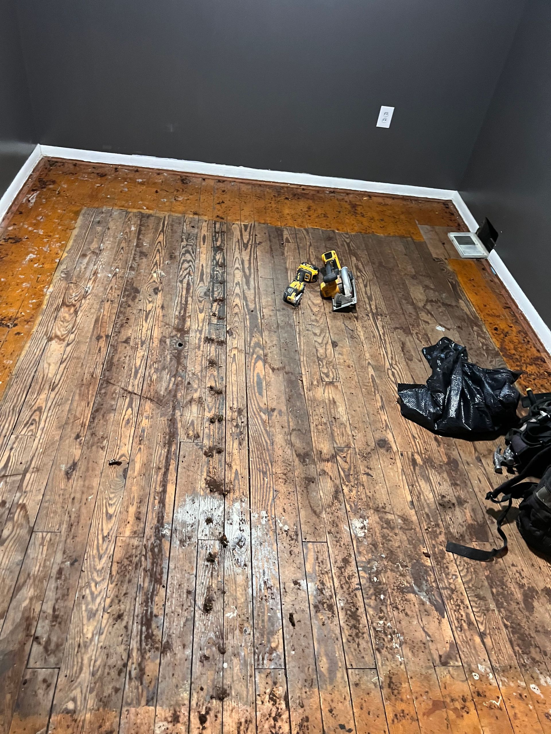 Room with torn-up wood flooring, tools, and debris. White trim around the perimeter, black ceiling, dark walls.
