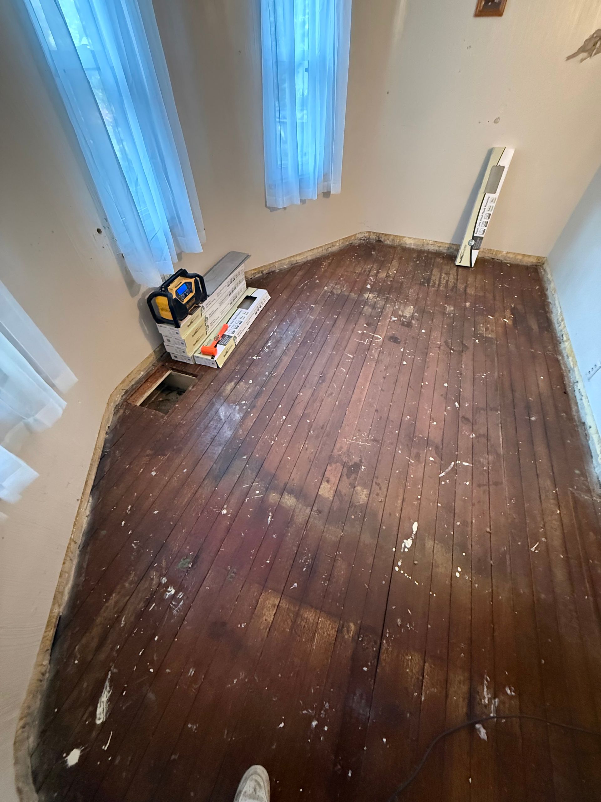 Room with exposed hardwood floors, tools, and two windows. Walls are beige, light blue curtains.