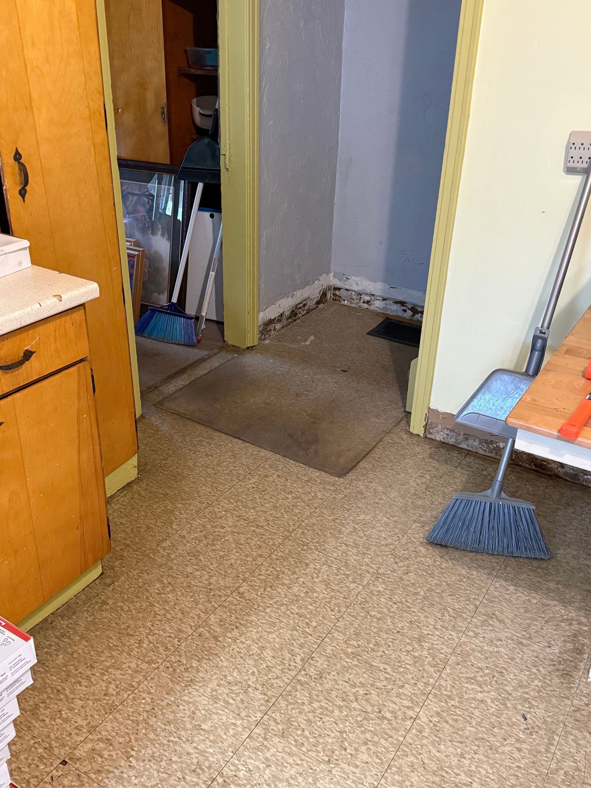 Kitchen with linoleum floor, cabinets, broom. Doorway leads to a second room.