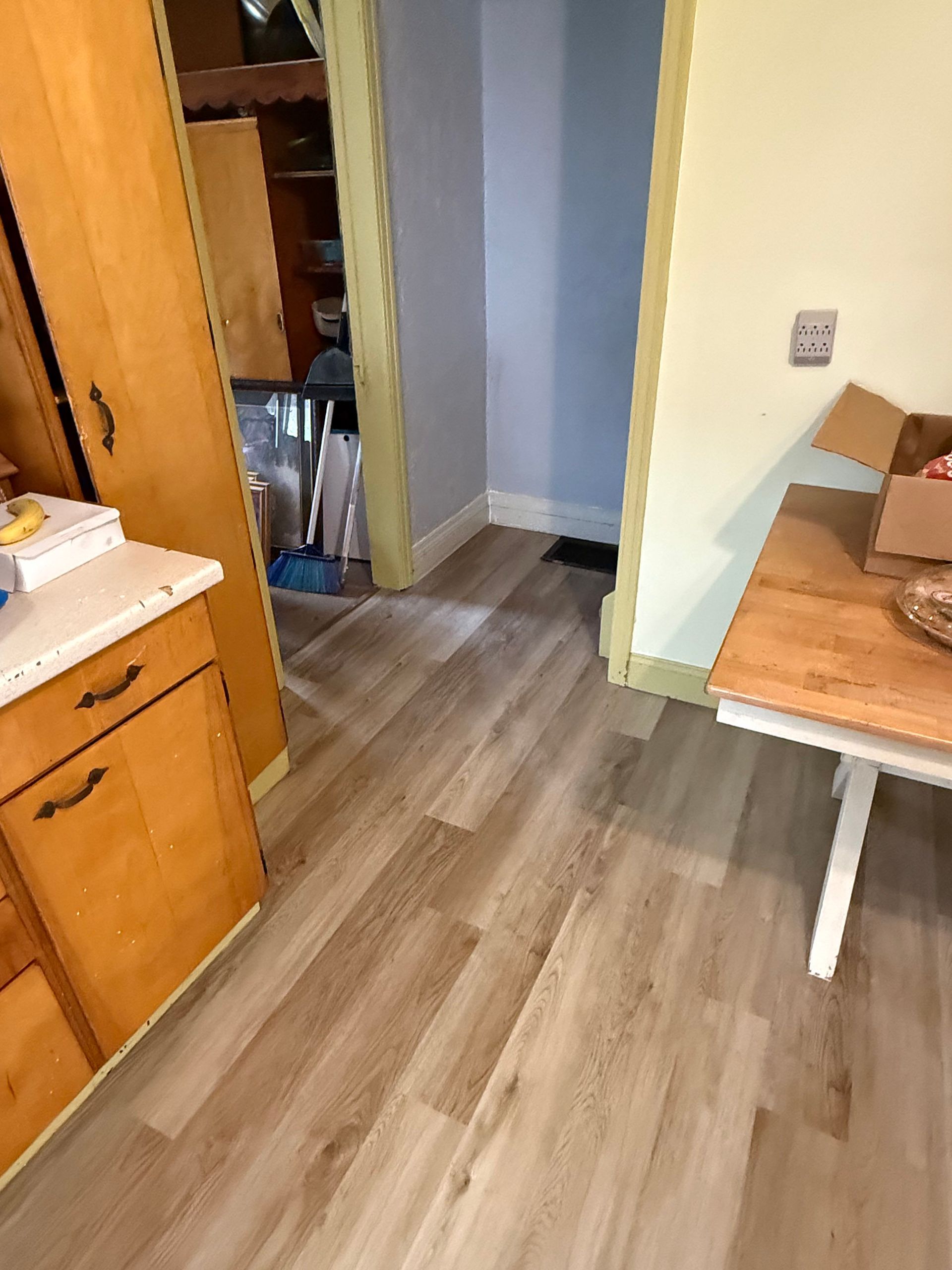 Kitchen with light wood-look flooring, light yellow walls, and a view into a hallway. A cabinet and table are visible.
