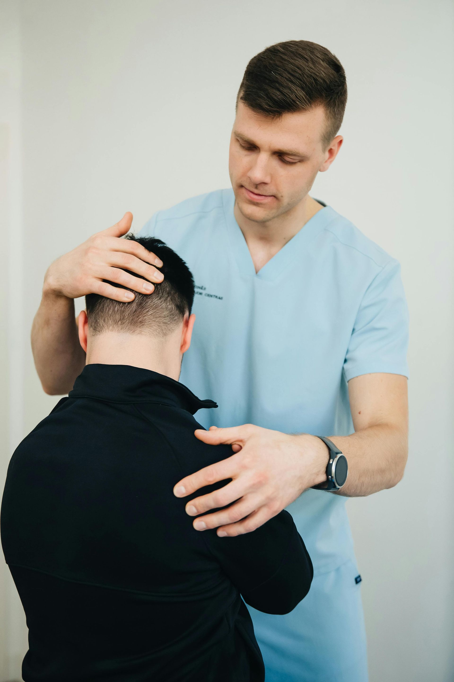 A doctor in blue scrubs examining a patient's head and shoulders in a light-colored room.