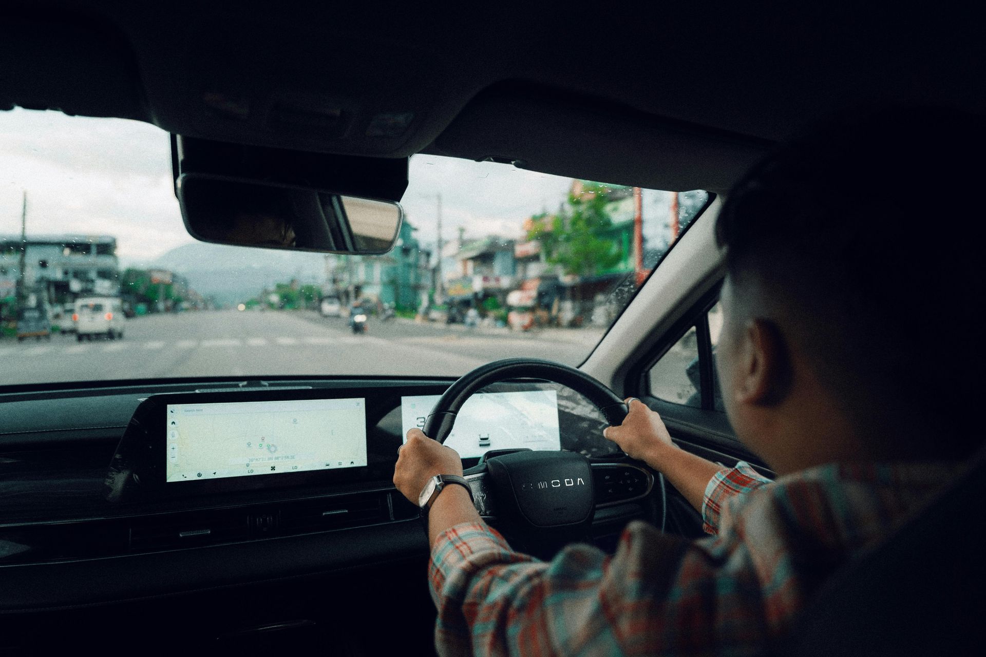 Person driving a car, hands on the steering wheel. Interior view with road and buildings visible through windshield.
