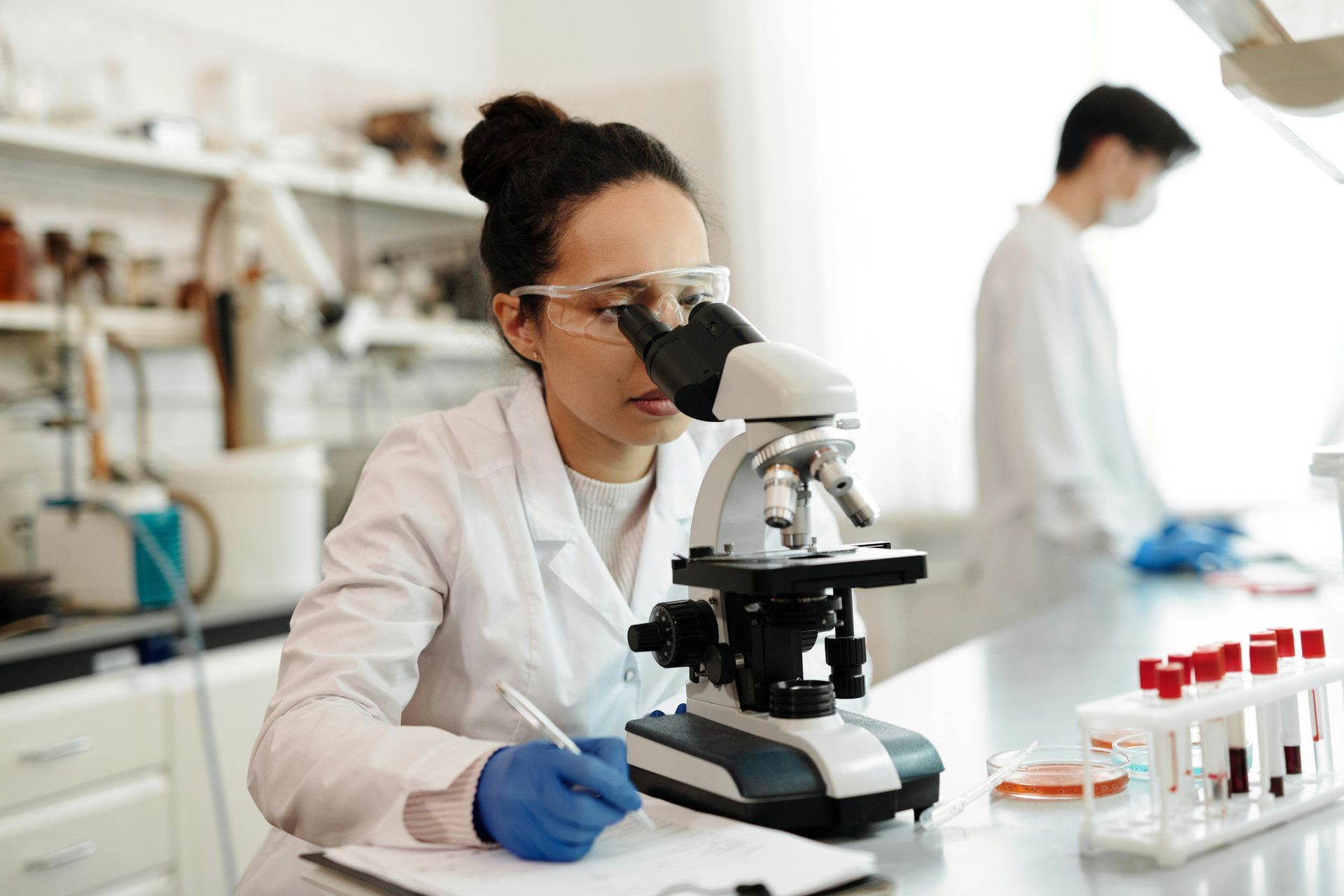 Woman in lab coat using microscope, writing notes. Another person in background, vials on table.