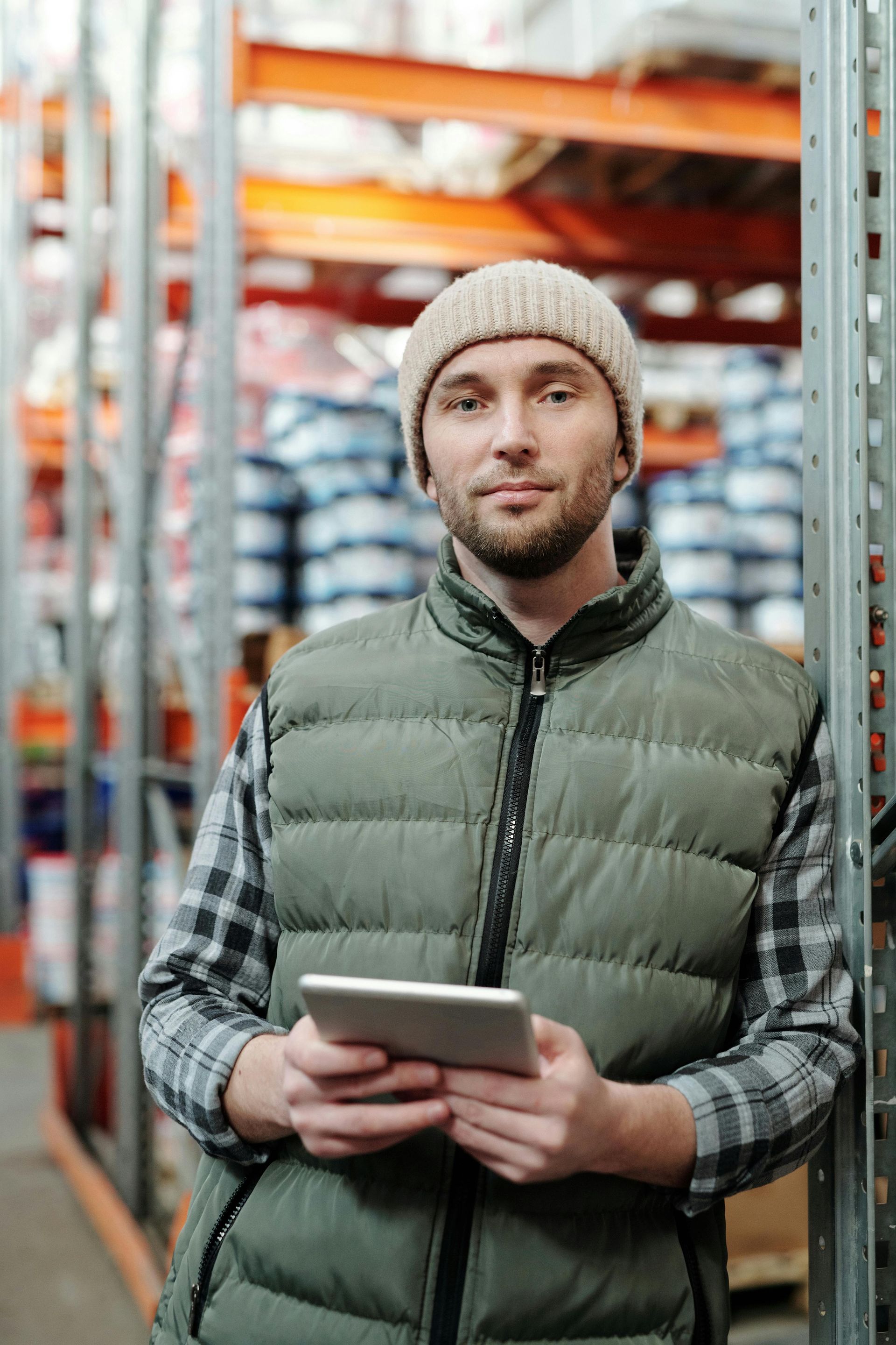 Warehouse worker in beanie and vest holding tablet, smiling, leaning on shelving.