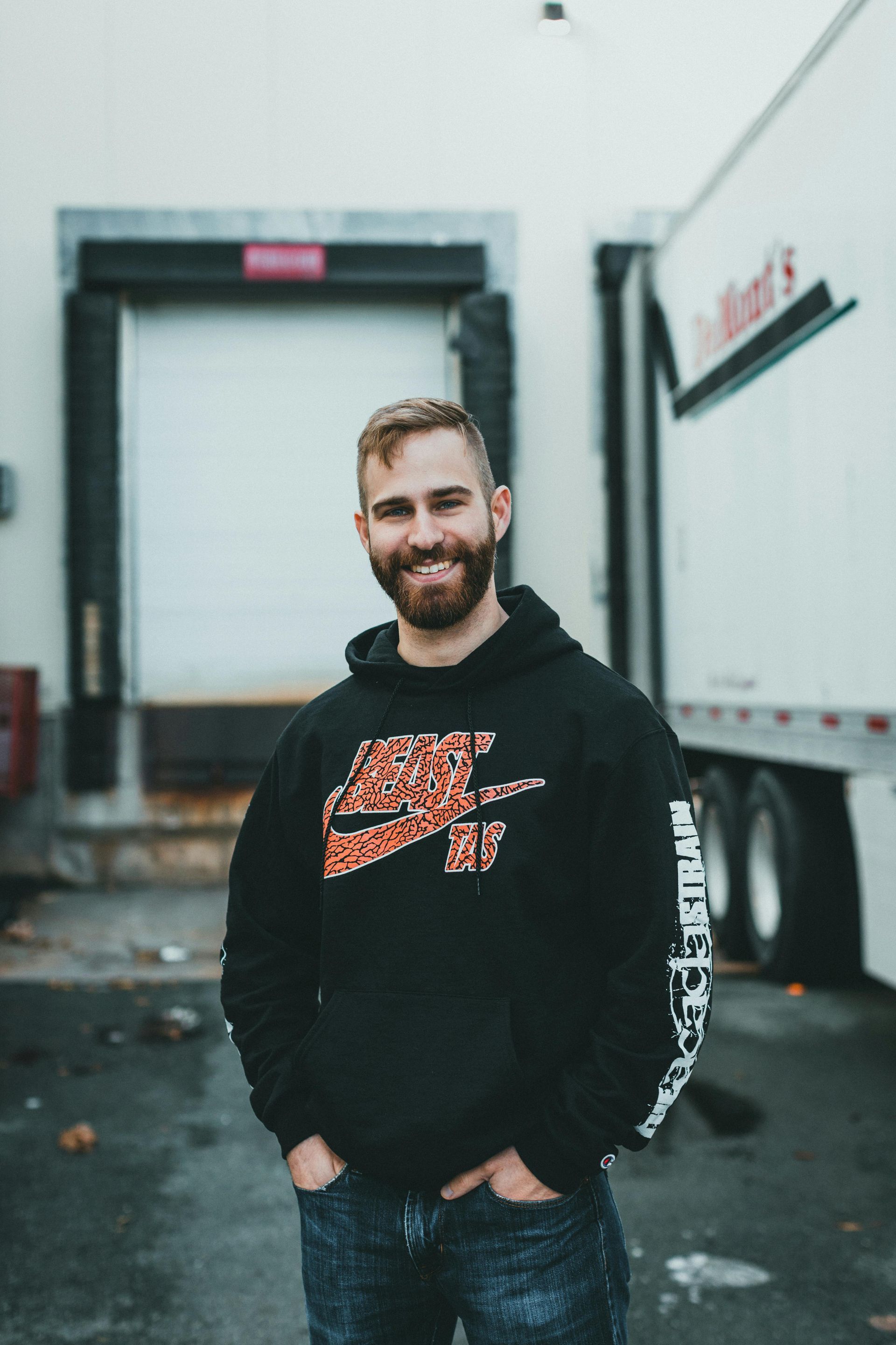 Man in black Nike hoodie smiles in front of a loading dock and semi-trailer.