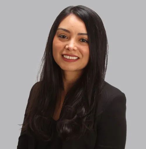 Woman with dark hair smiles, wearing a black blazer against a gray background.