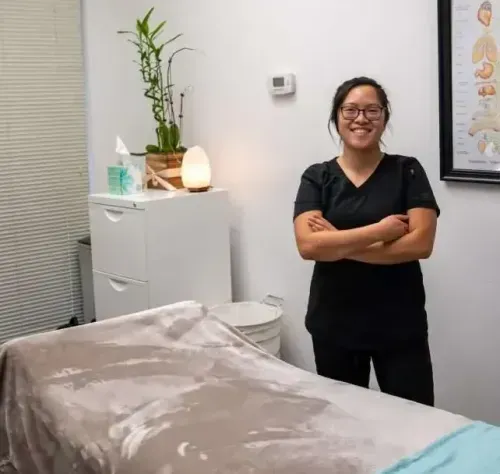 Woman in black scrubs stands in a spa room, arms crossed, smiling. Massage table in foreground.