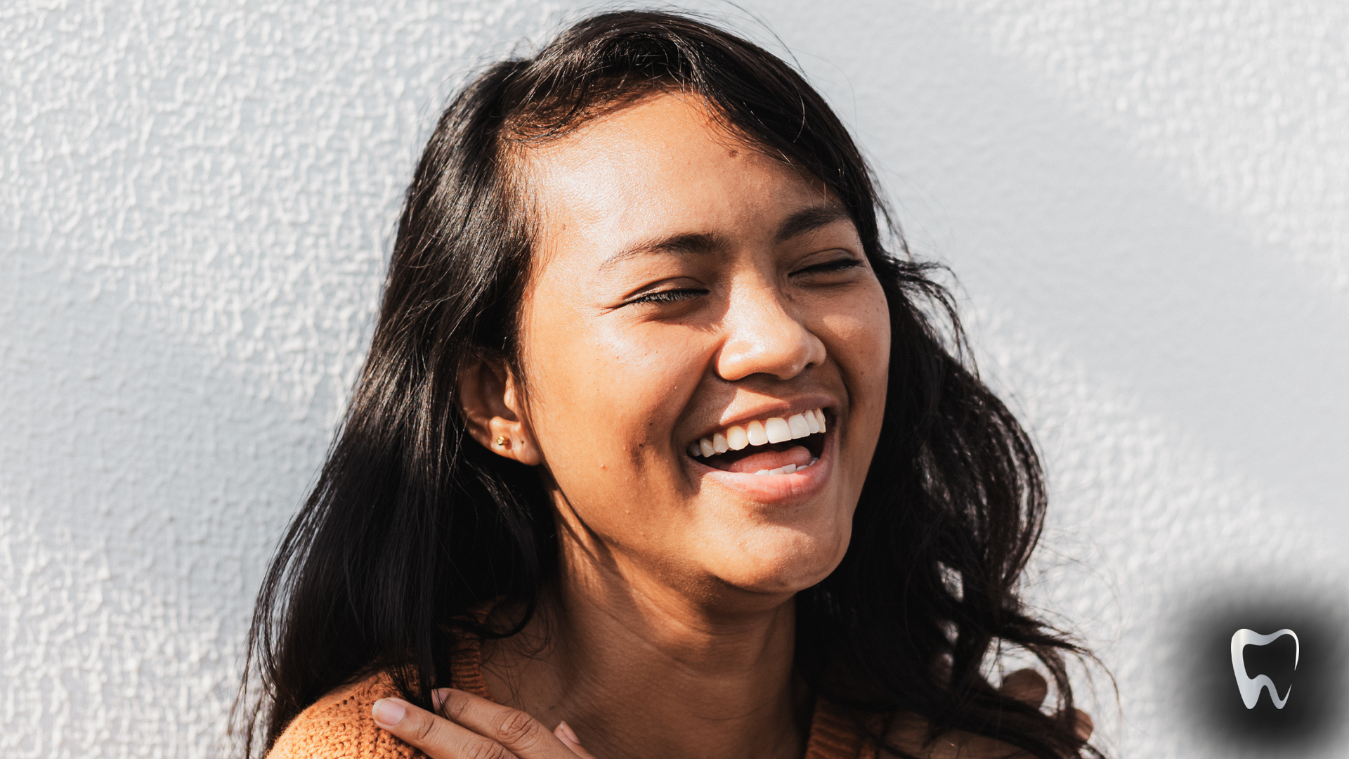 Woman with long dark hair laughs, leans against a white wall.
