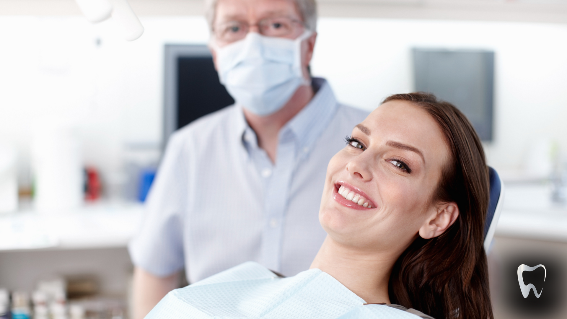 A woman is smiling while sitting in a dental chair with a dentist in the background.