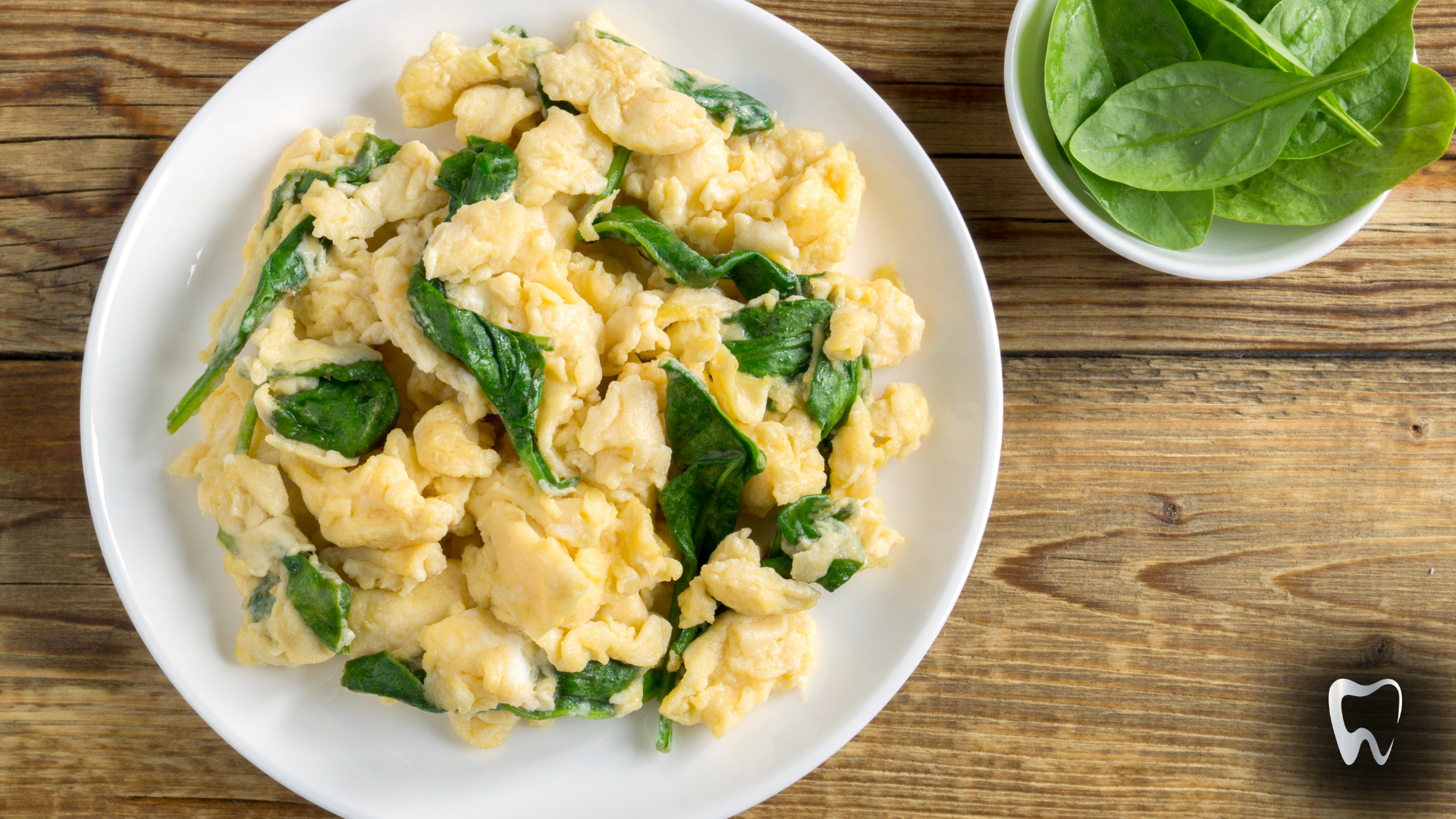 A plate of scrambled eggs with spinach on a wooden table next to a bowl of spinach.