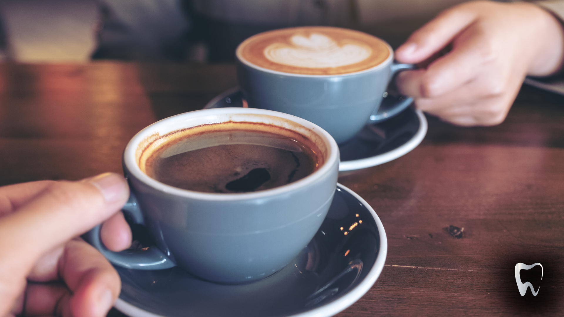 Two people holding coffee cups on a wooden table. One cup has a latte, the other is dark.