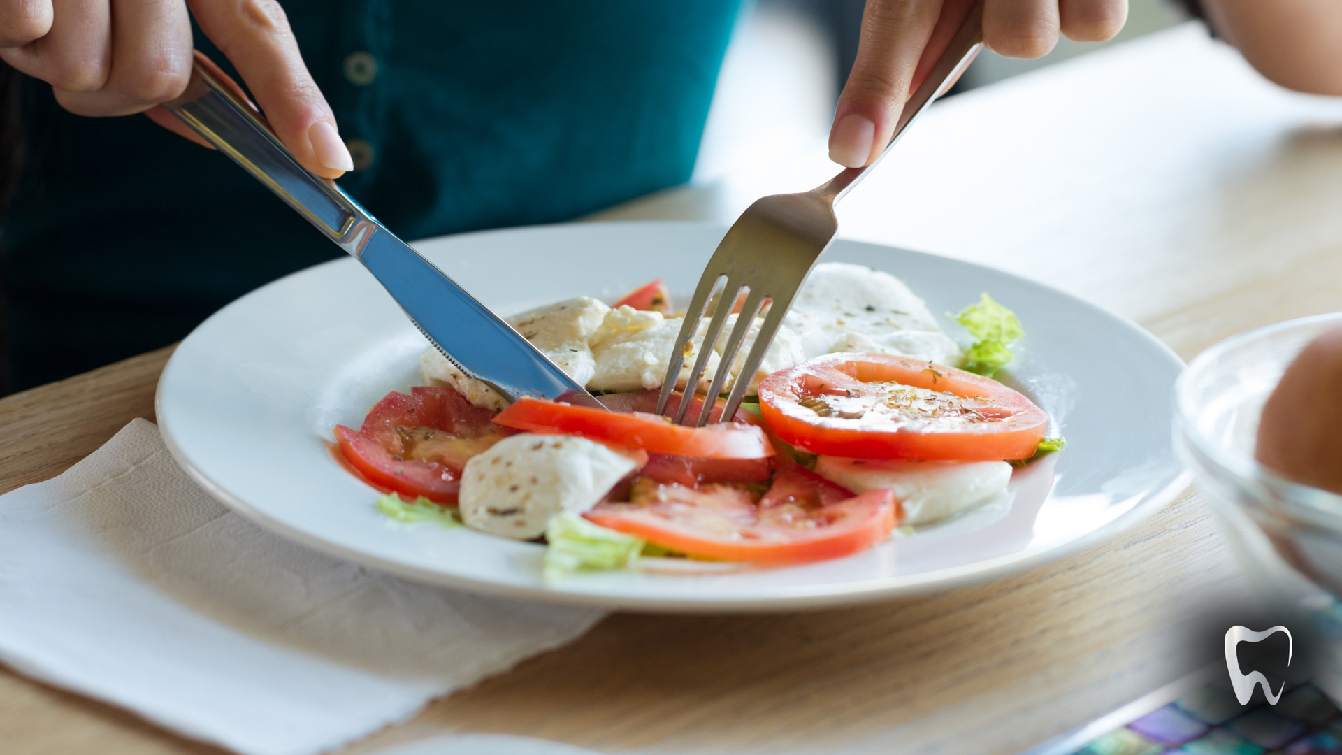 Woman cutting a tomato and mozzarella salad with a knife and fork on a white plate.