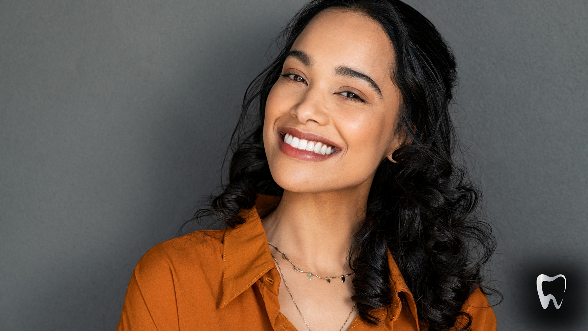 A woman in an orange shirt is smiling and wearing a necklace.