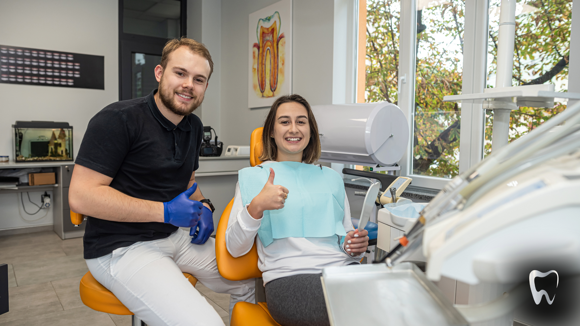 A man and a woman are sitting in a dental chair and the woman is giving a thumbs up.