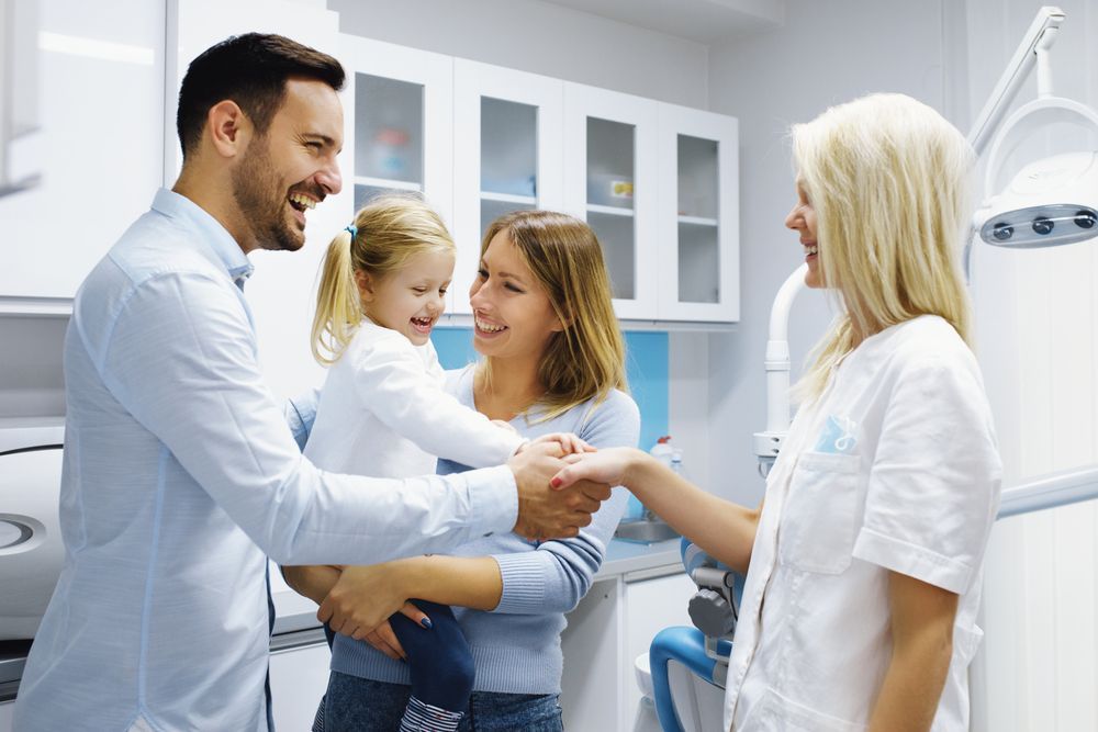 Family smiling and giving thumbs-up in a well-lit room with white cabinets; the woman holds a child.