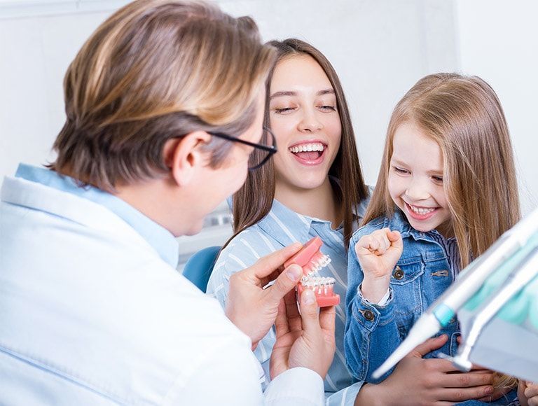 A dentist is showing a woman and a little girl how to brush their teeth.