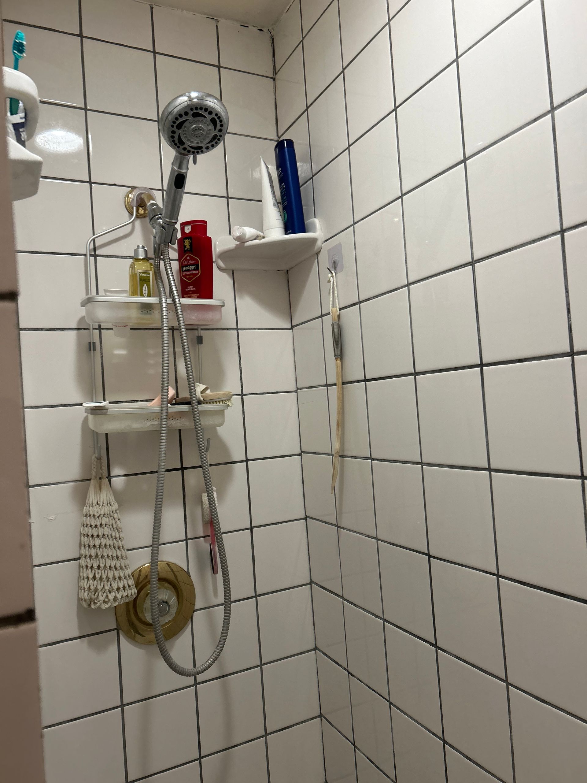 Small white tiled shower with showerhead, shelves holding toiletries, and a hanging loofah.